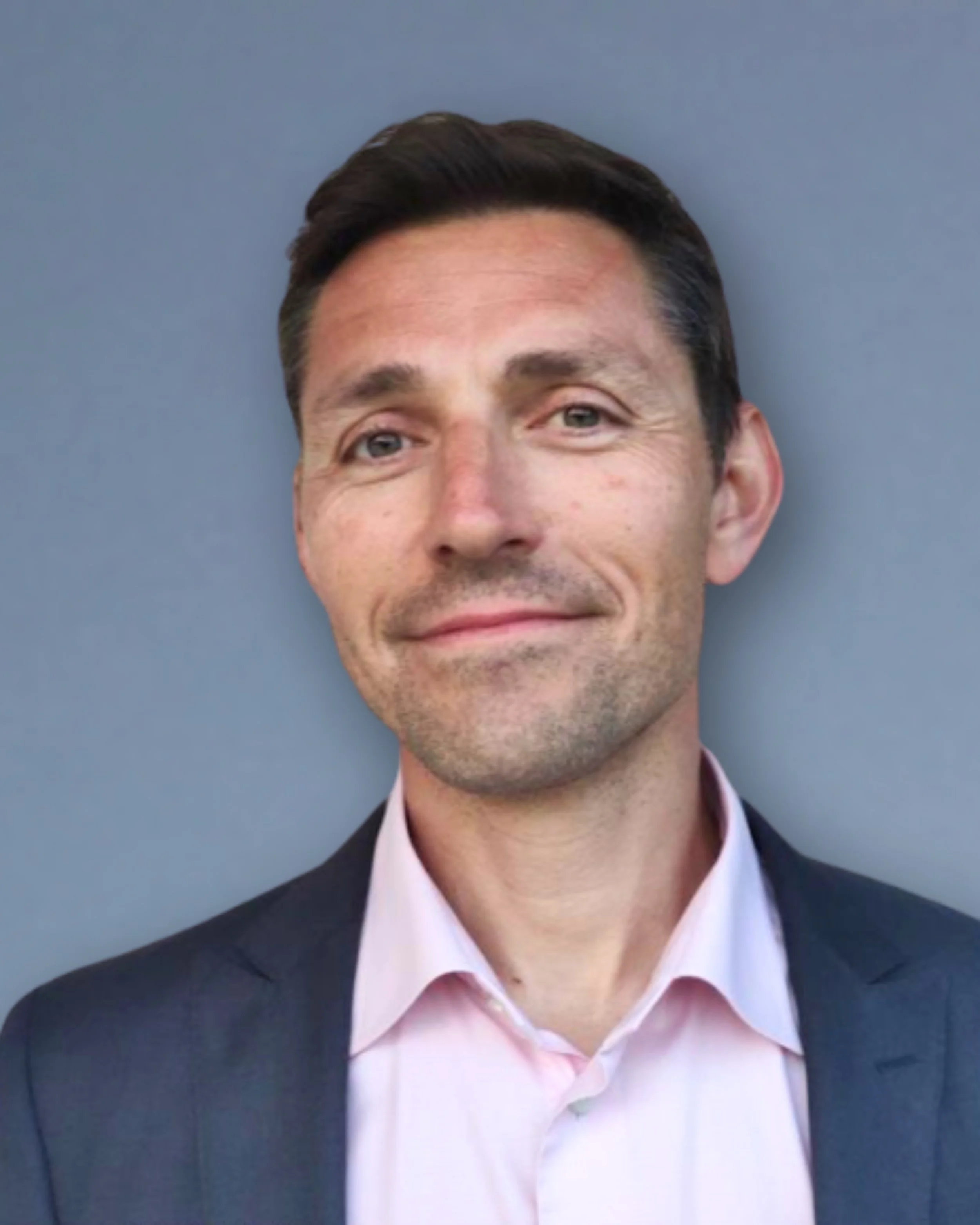 A man in a suit with a pink shirt, smiling against a gray background.