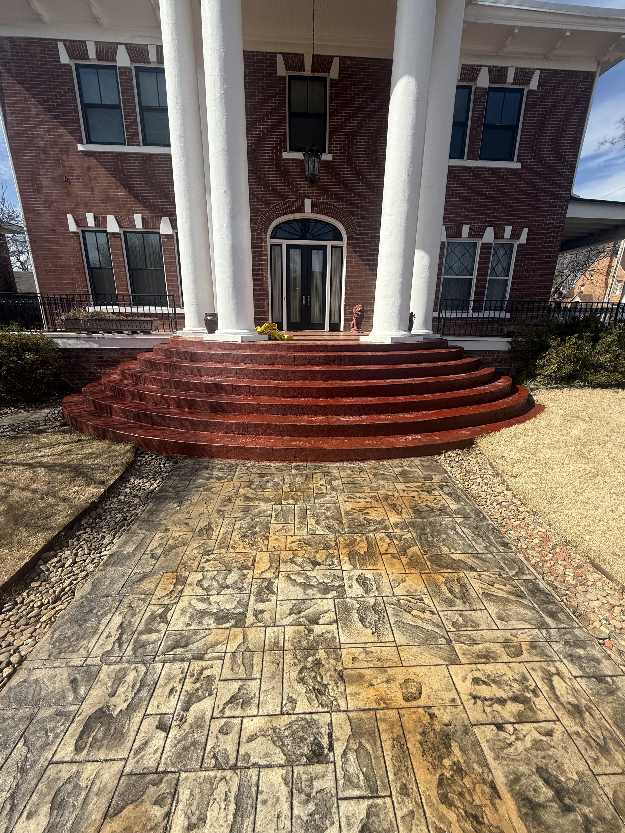 Front porch of a brick house with a round red brick staircase leading up to a black door, supported by white columns, with a patterned stone pathway in the front yard.