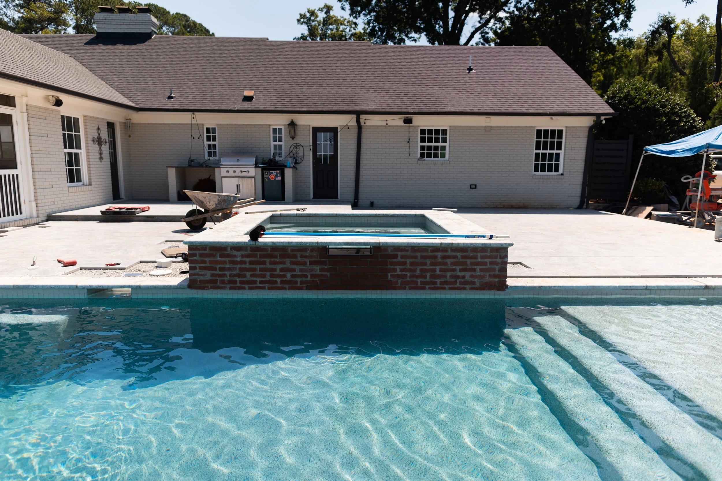 Backyard with a swimming pool under construction, including a partially built hot tub, construction tools, and outdoor furniture. A single-story house with a gray roof and white brick walls is in the background.