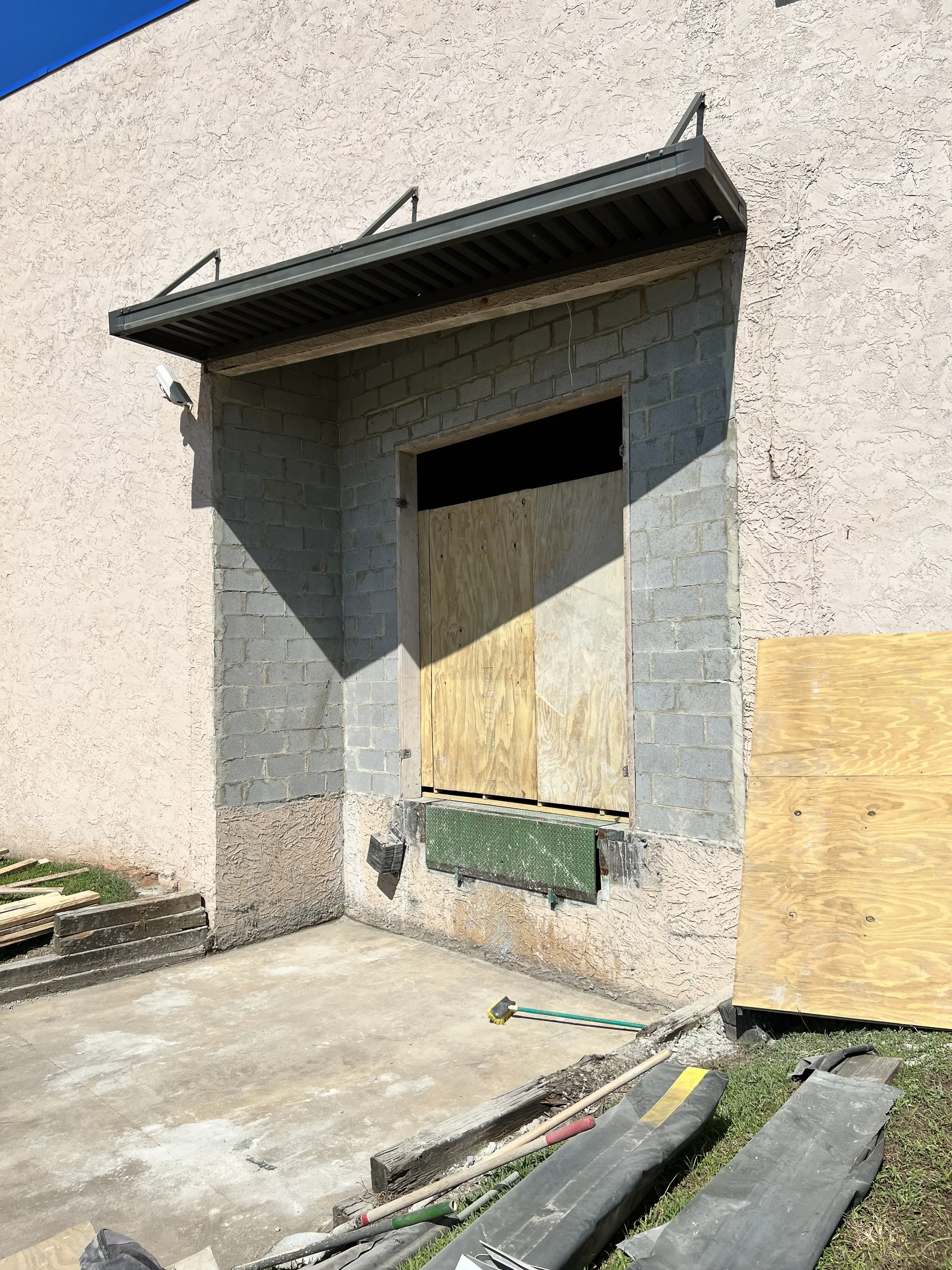 Building under construction with a boarded-up window, a small overhang, and construction tools on the ground.