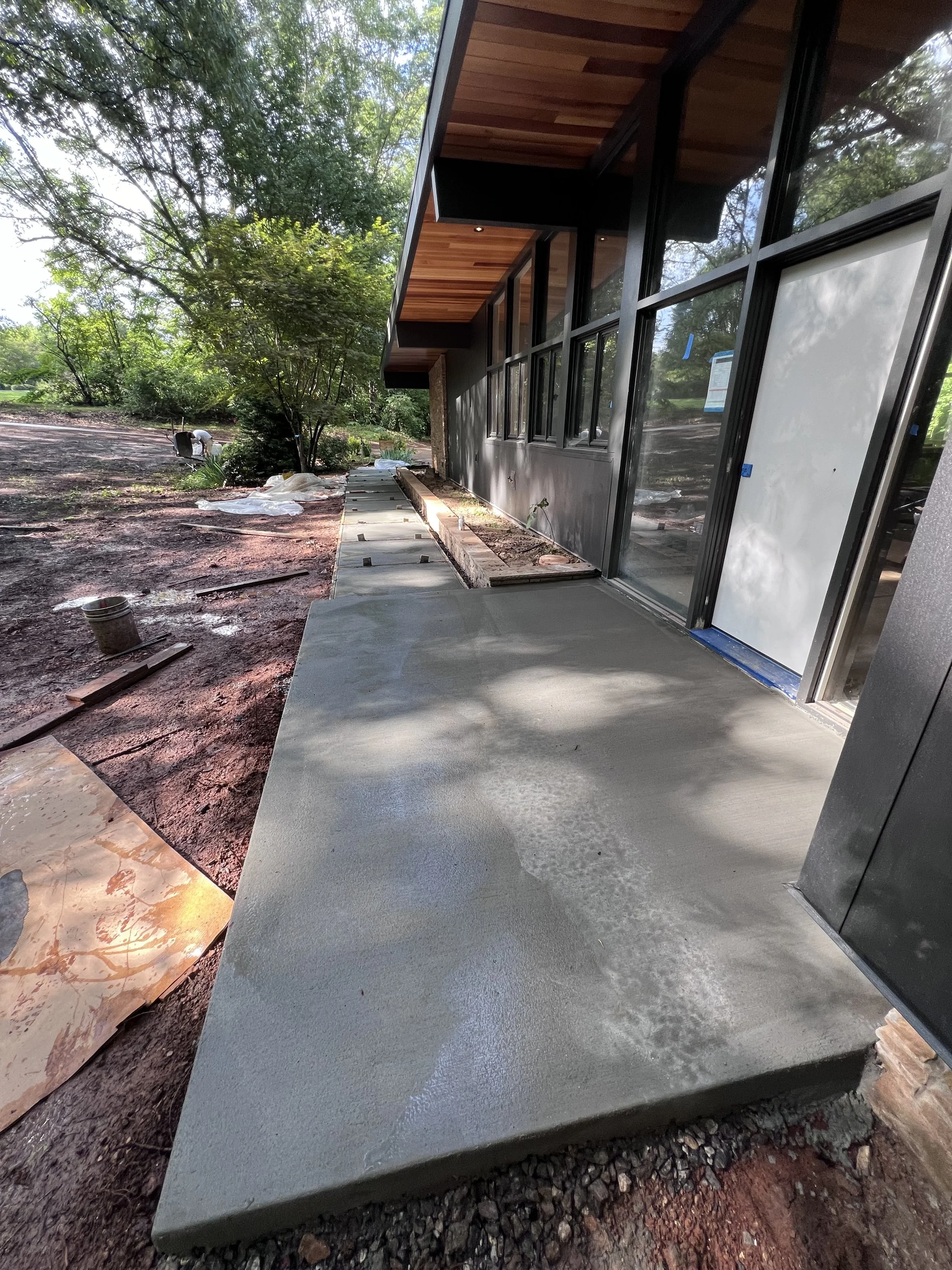 Newly poured concrete patio next to a modern house with large glass windows, surrounded by trees on a sunny day.