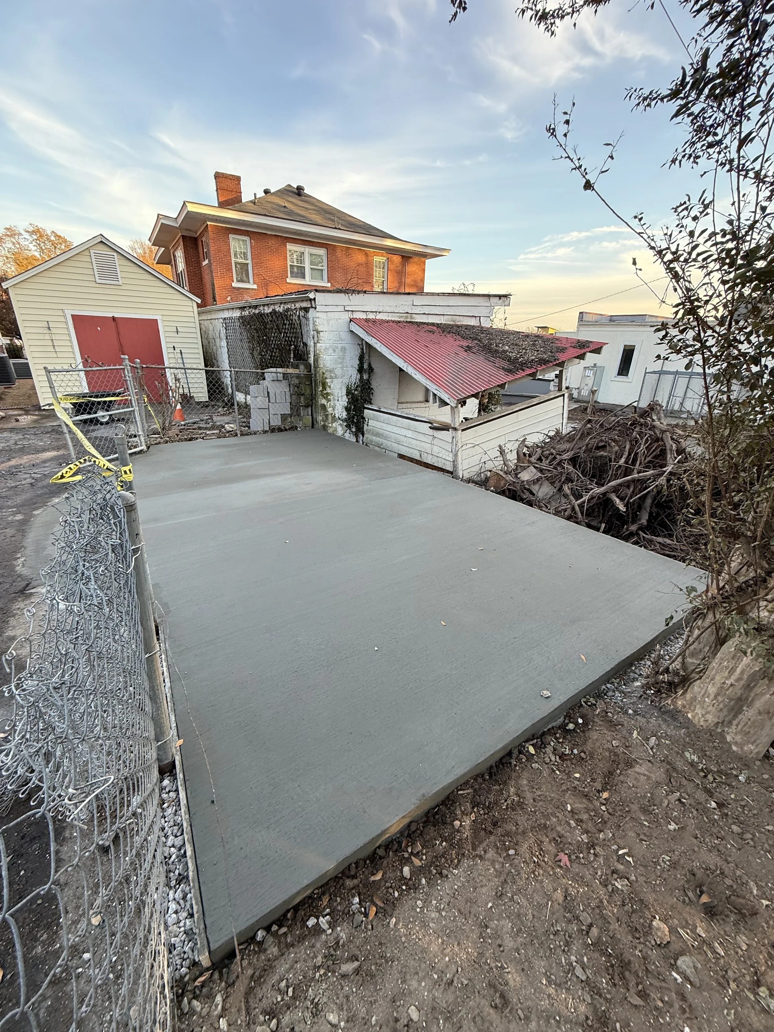 Freshly poured concrete slab in an outdoor backyard, surrounded by construction fencing and a chain-link fence.