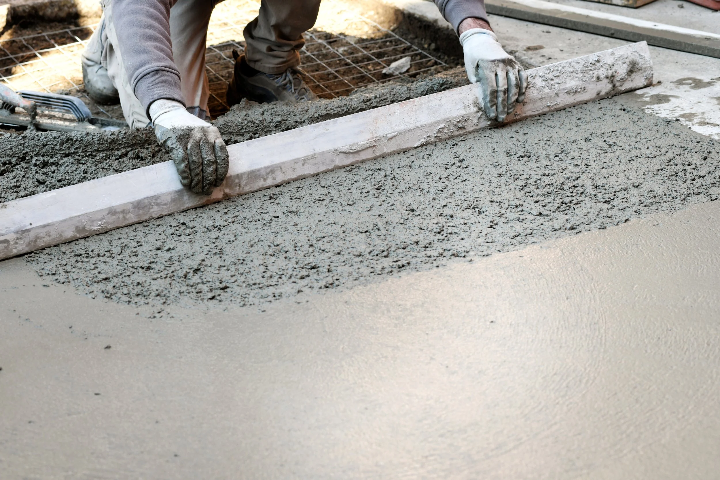 A construction worker is smoothing wet concrete with a straightedge tool during a concrete pouring process.