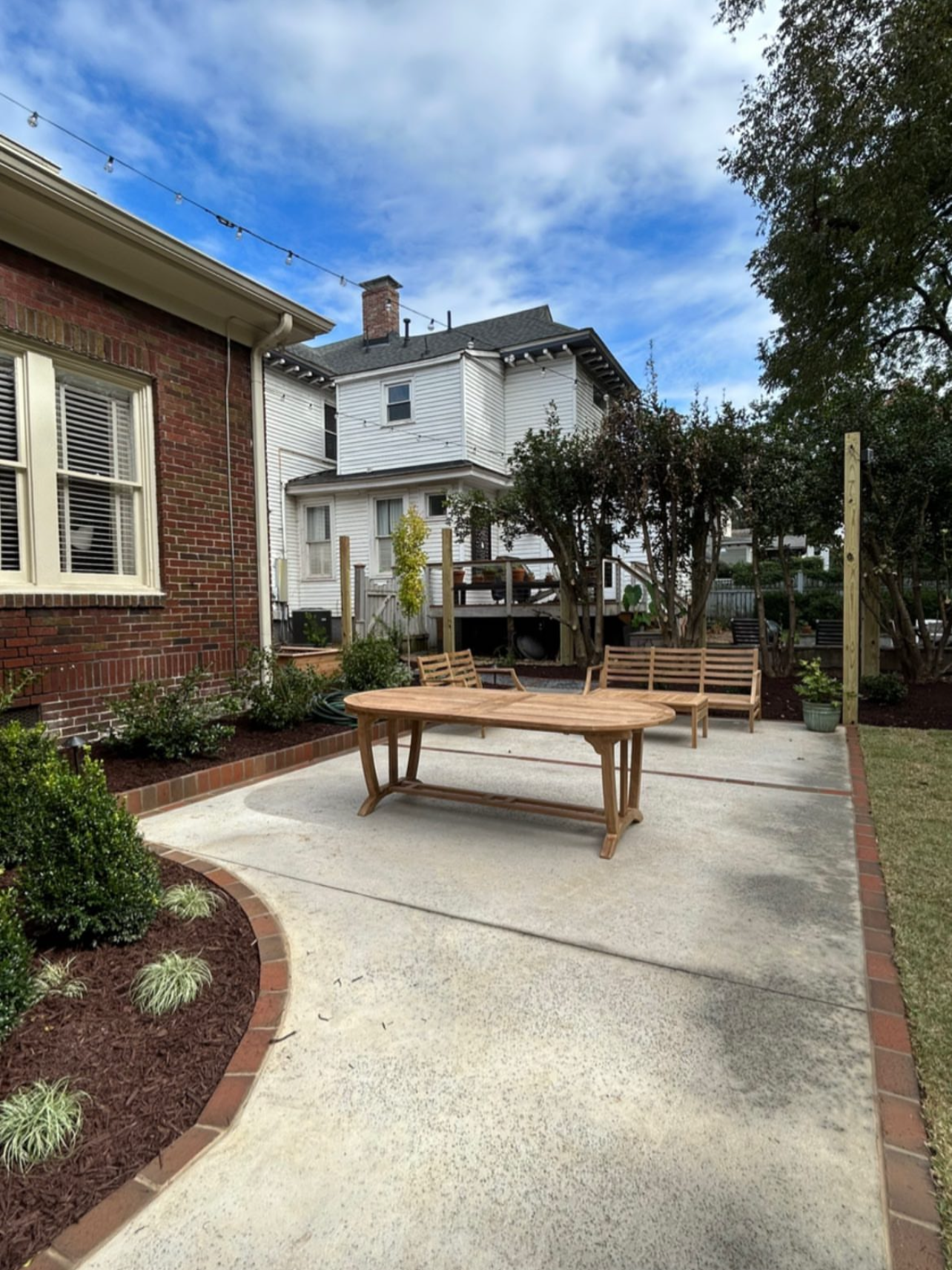 Backyard patio with wooden furniture, including a table and benches, surrounded by landscaped garden beds, and backyard houses and trees in the background under a partly cloudy sky.
