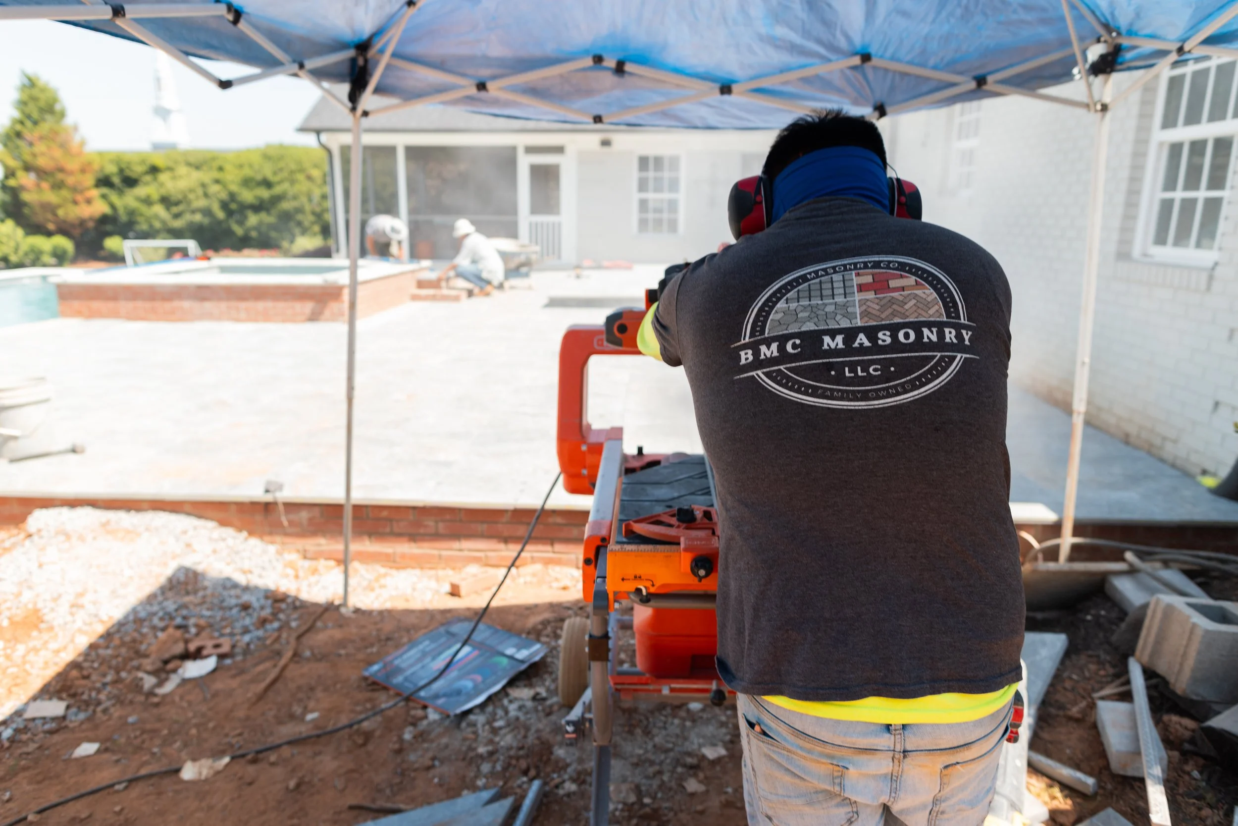 A construction worker saws wood outdoors under a blue canopy with a house in the background, and two individuals working near a pool in the distance.