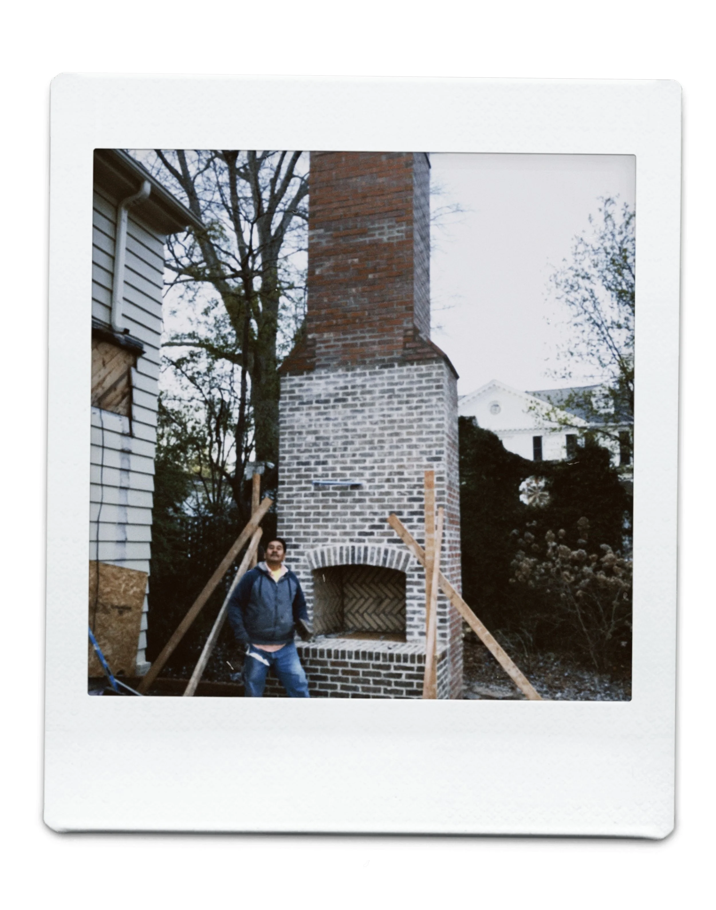A man standing in front of a new brick and chimney outdoor fireplace under construction, with wooden support beams around it.