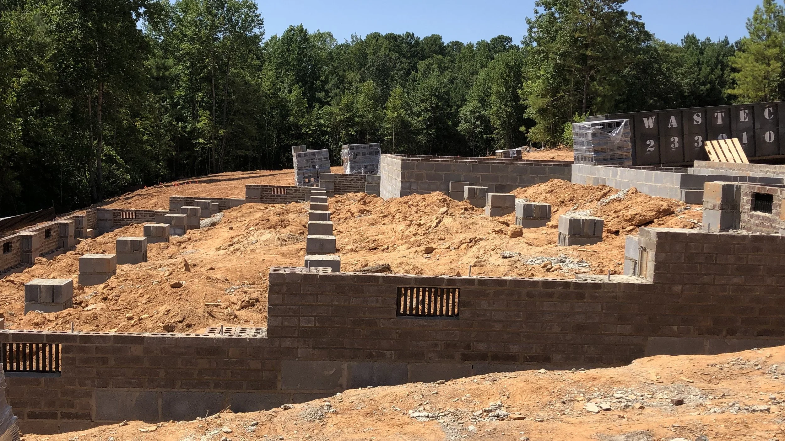Construction site with partially built brick walls and orange dirt, surrounded by trees.