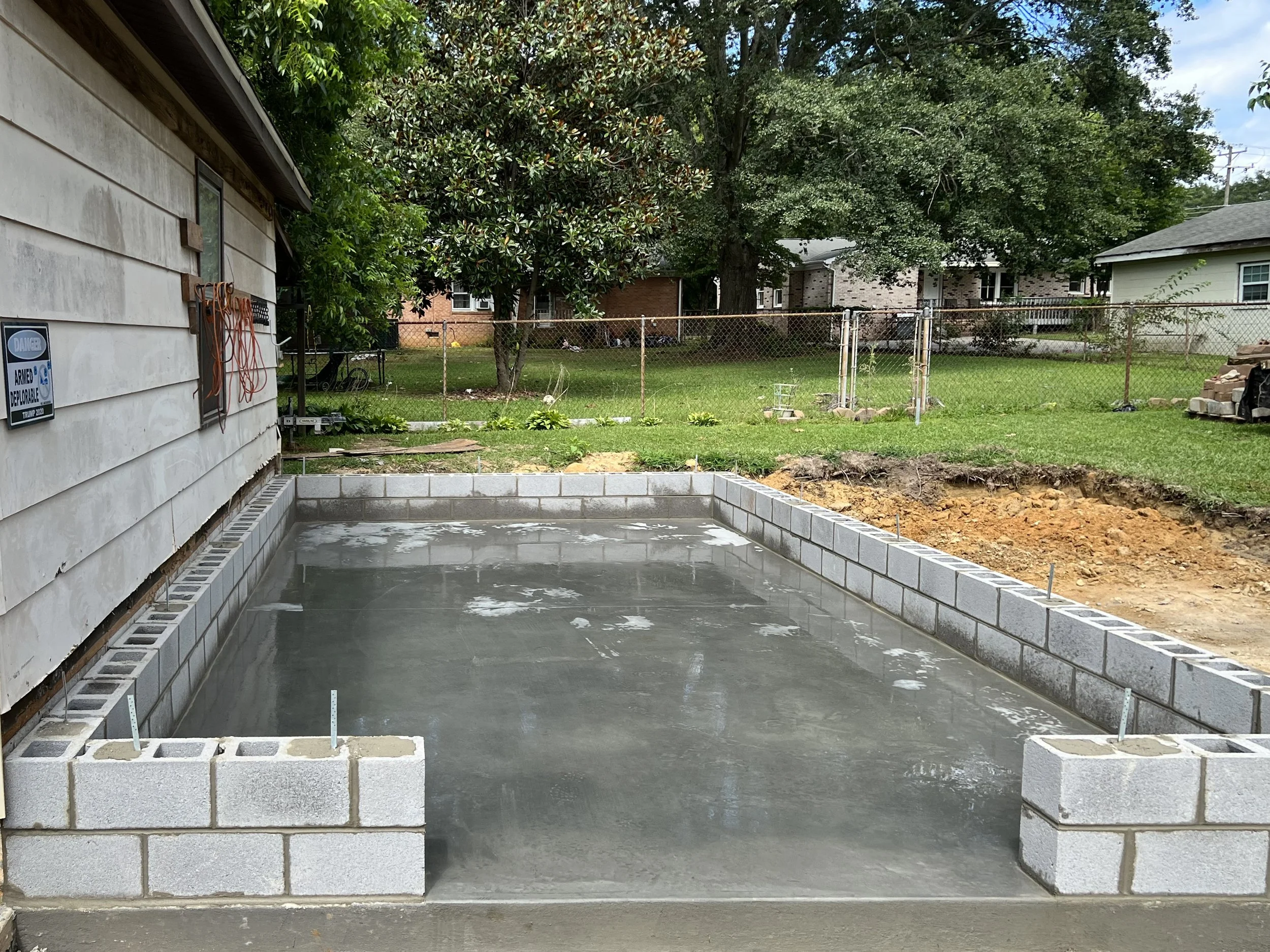 A poured concrete slab foundation with cinder block walls partially built around it, in a backyard under construction. The area is fenced and has trees and other houses in the background.