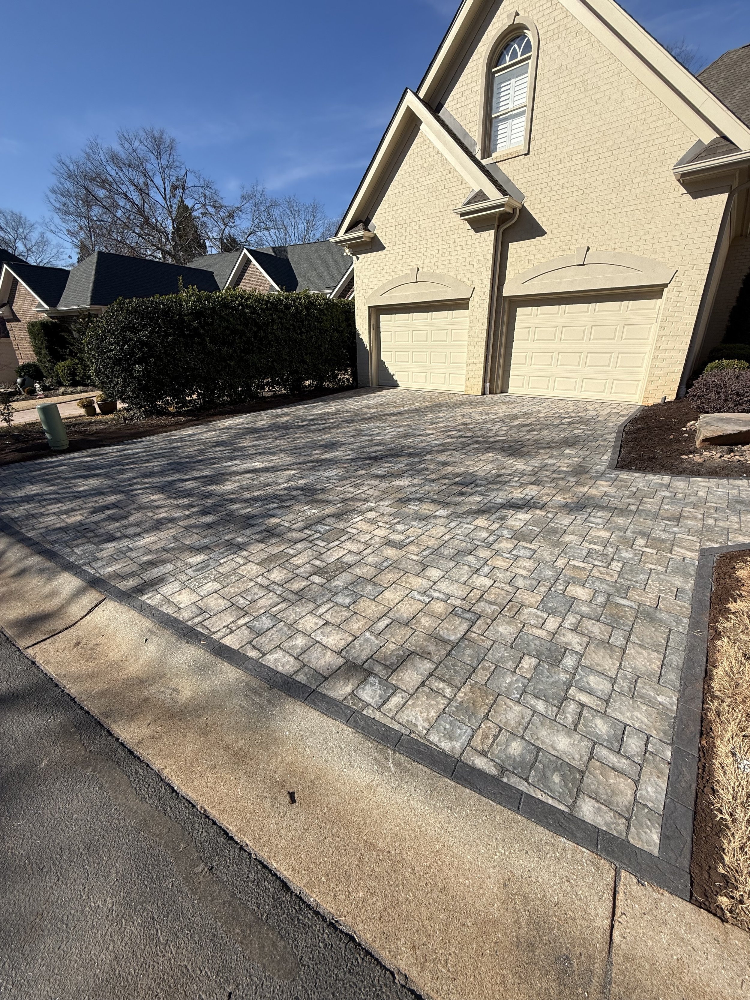 A newly paved stone driveway in front of a beige house with two garage doors and an arched window above. There are bushes on the left side and a flower bed with mulch on the right. The sky is clear with some trees in the background.