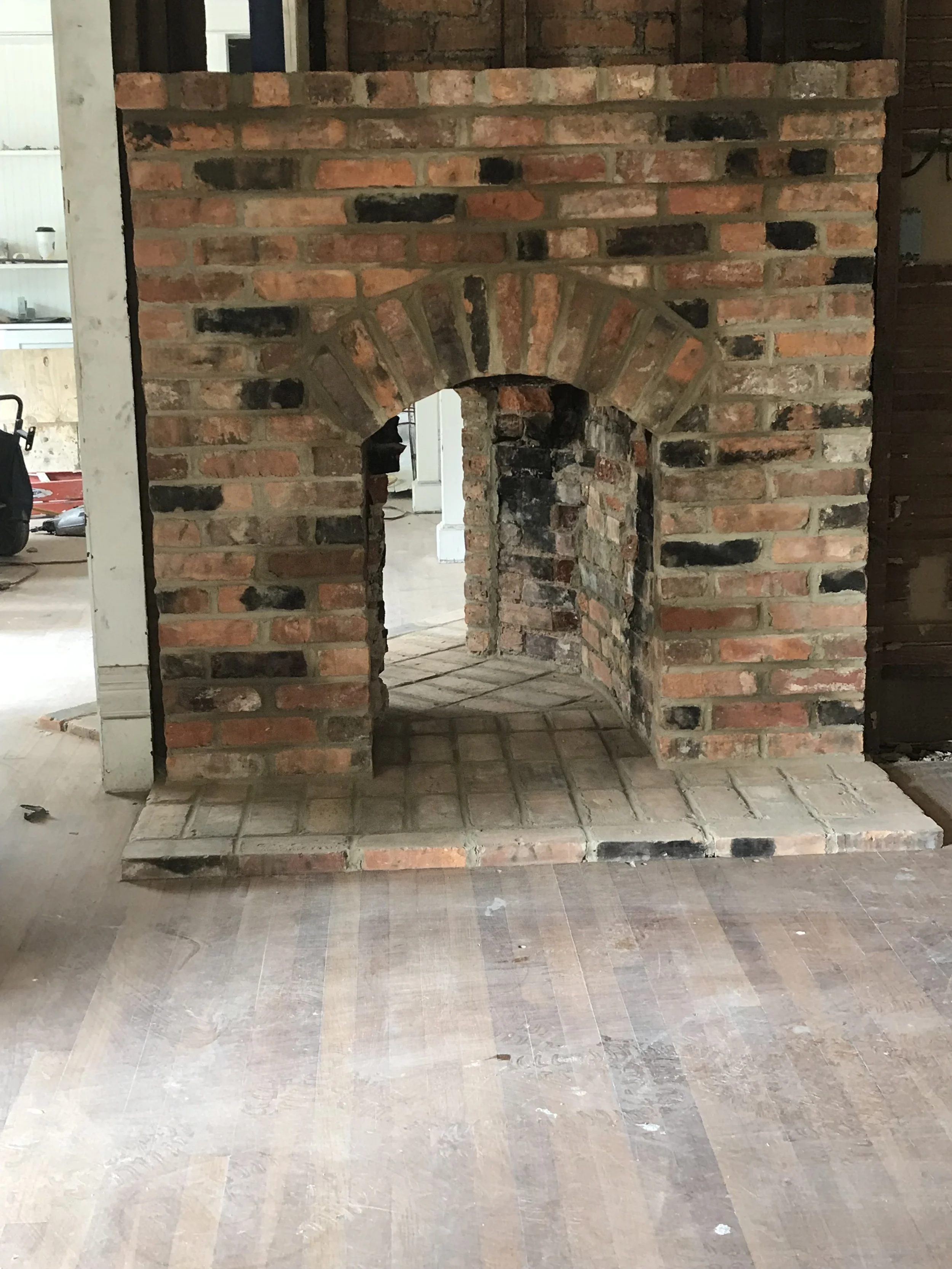 Unfinished brick fireplace with an arched opening inside a house under construction, with wooden floors and partially visible construction tools.