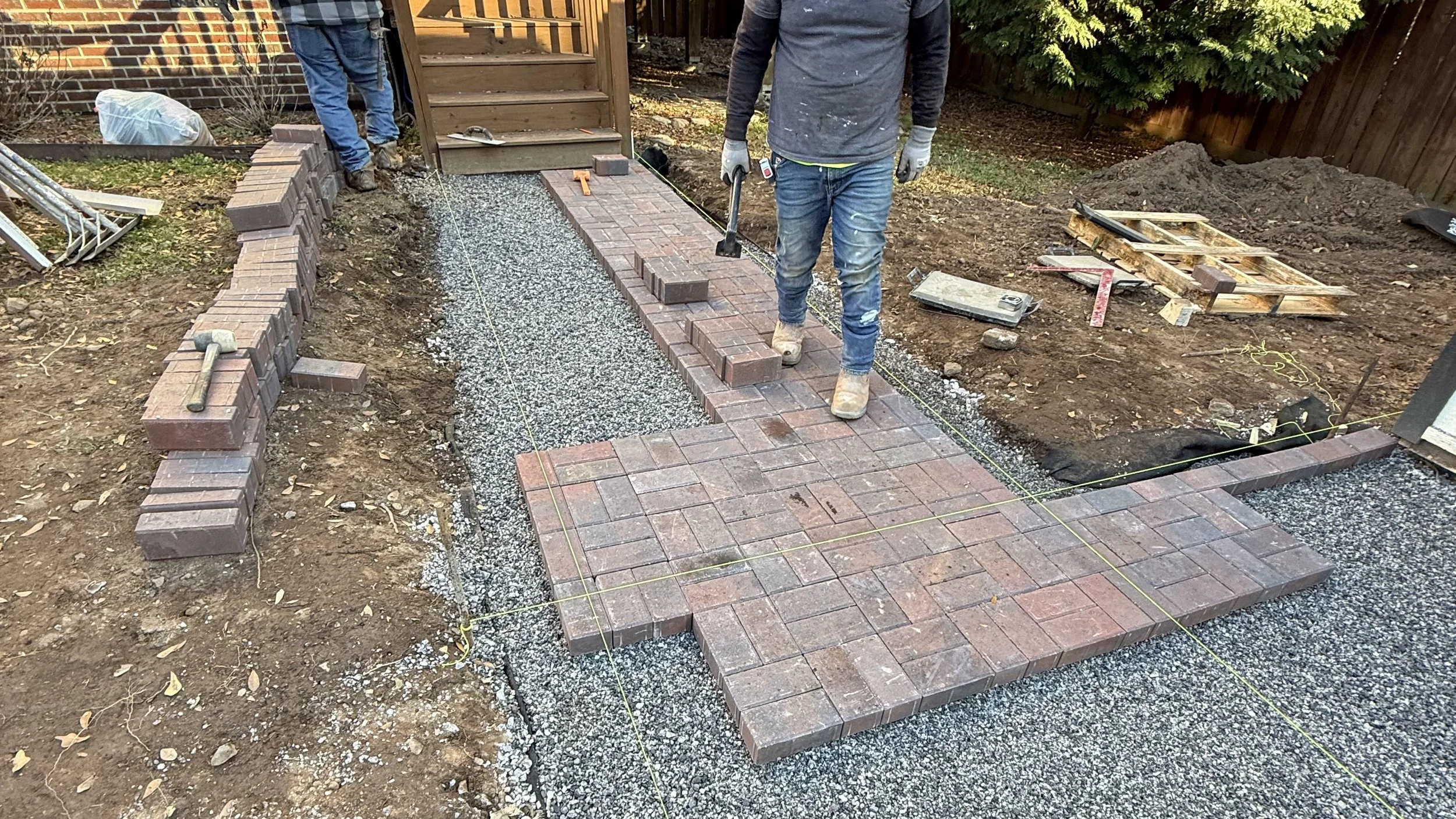 Workers constructing a brick pathway in a backyard with tools and materials, including bricks, gravel, and string guides.