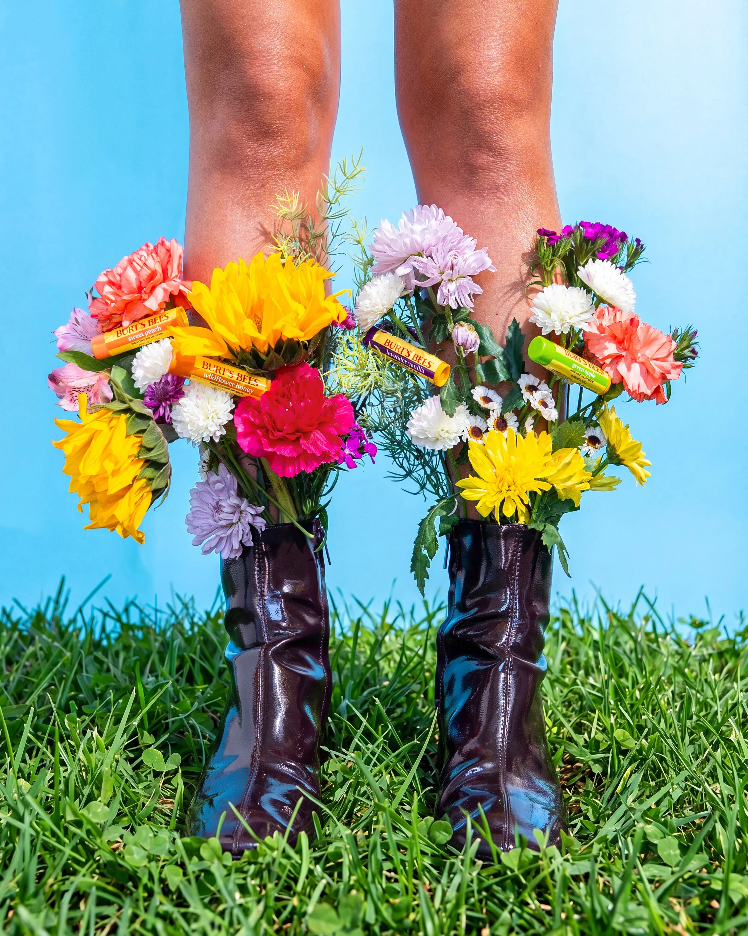Close-up of shiny black rain boots on grass with colorful flowers and Burt's Bees lip balm tubes tucked inside the flowers, against a bright blue background.