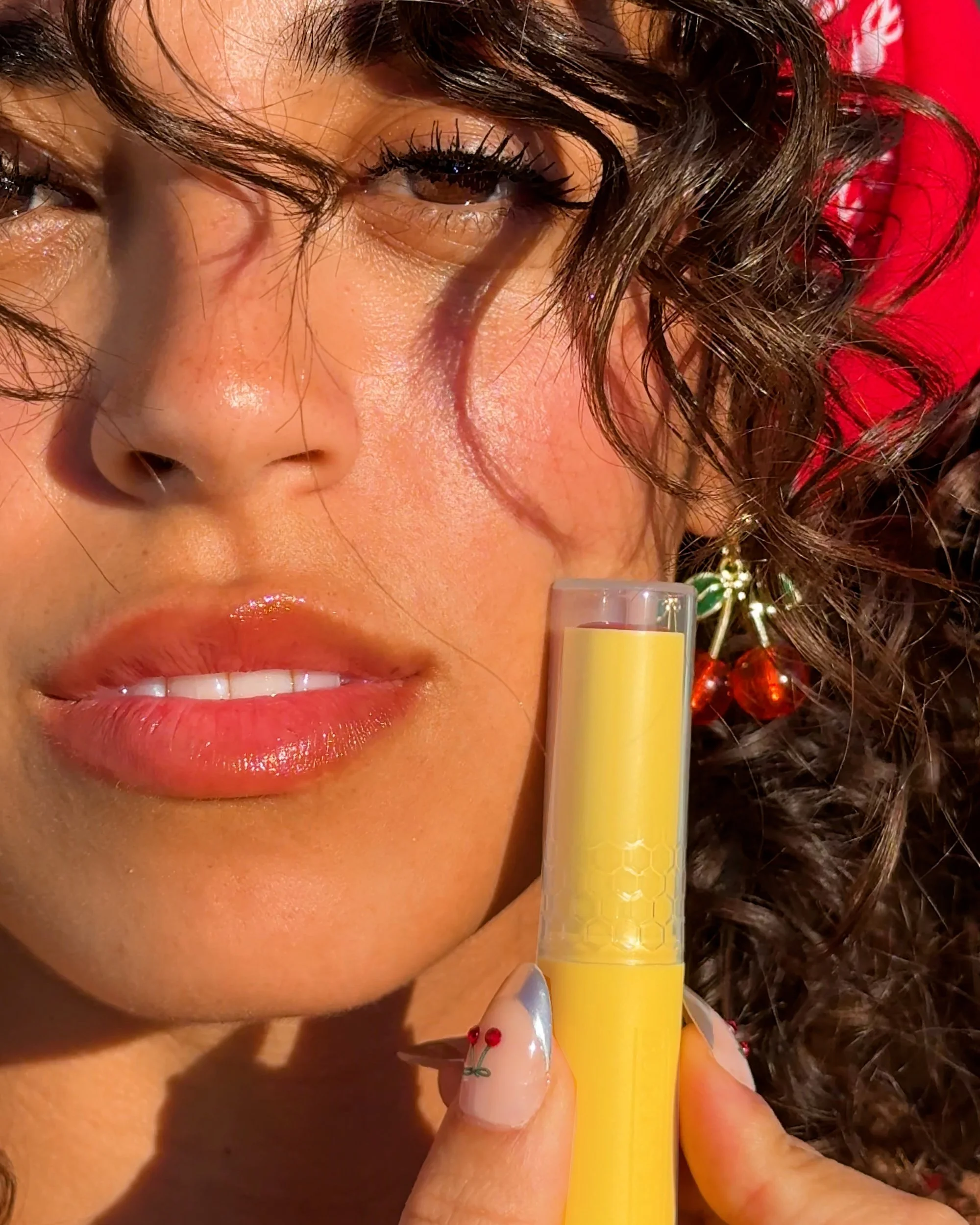 Close-up of a woman with curly hair holding a yellow tube of lip balm near her lips.