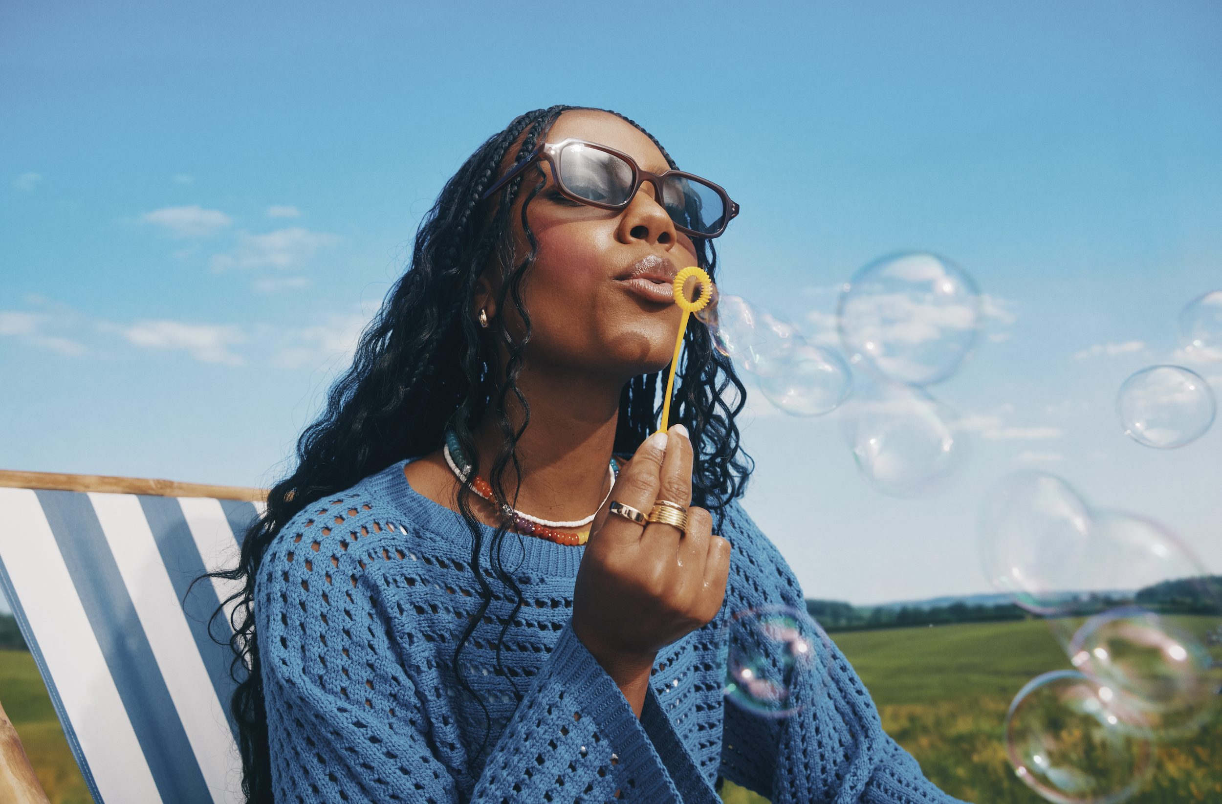 A woman with dark curly hair wearing sunglasses and a blue crochet sweater, blowing bubbles on a sunny day outdoors.