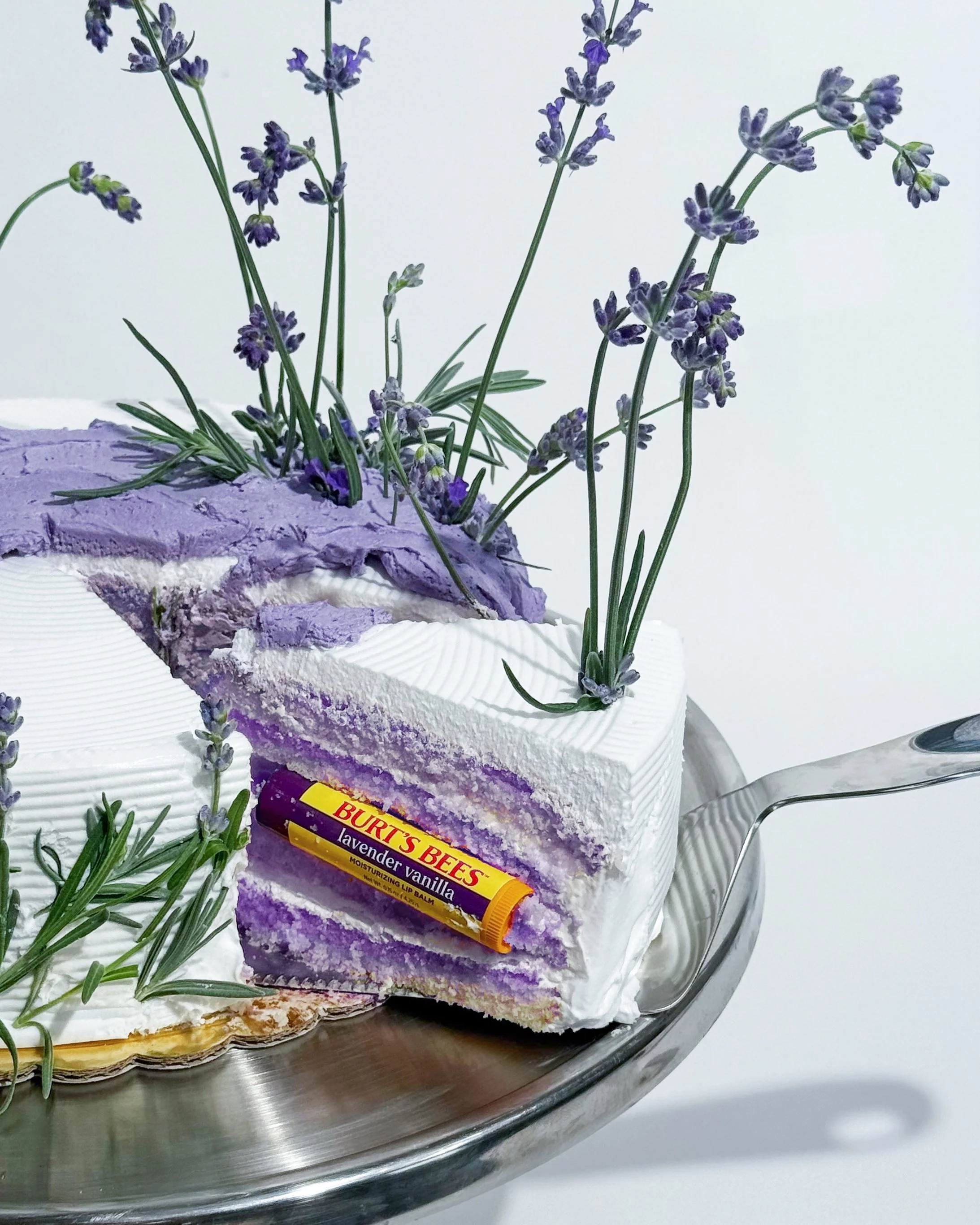 A cake decorated with purple sprigs of lavender, some placed on top and some on the side, with a slice showing purple and white layers and a lavender vanilla flavored Burt's Bees lip balm on top.
