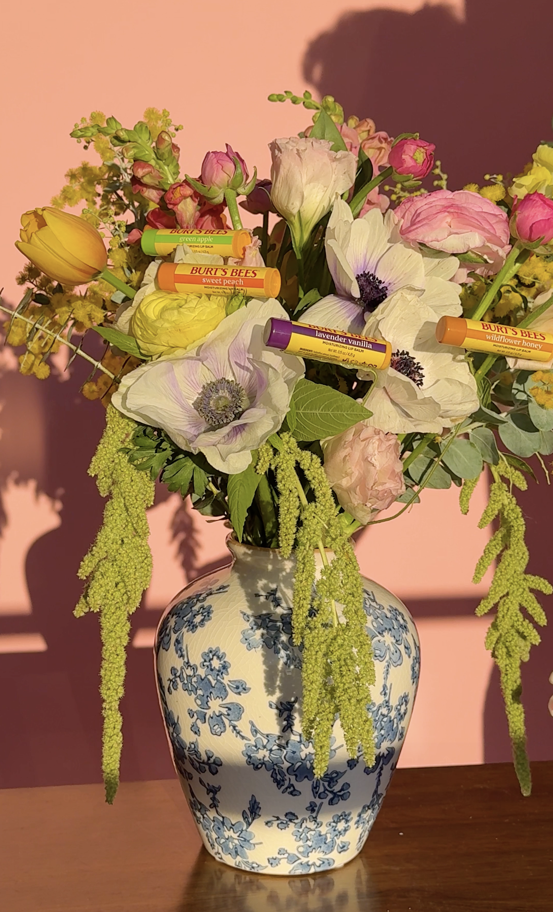 A floral arrangement in a blue and white patterned vase containing various flowers and lip balm sticks, with a pink wall background.