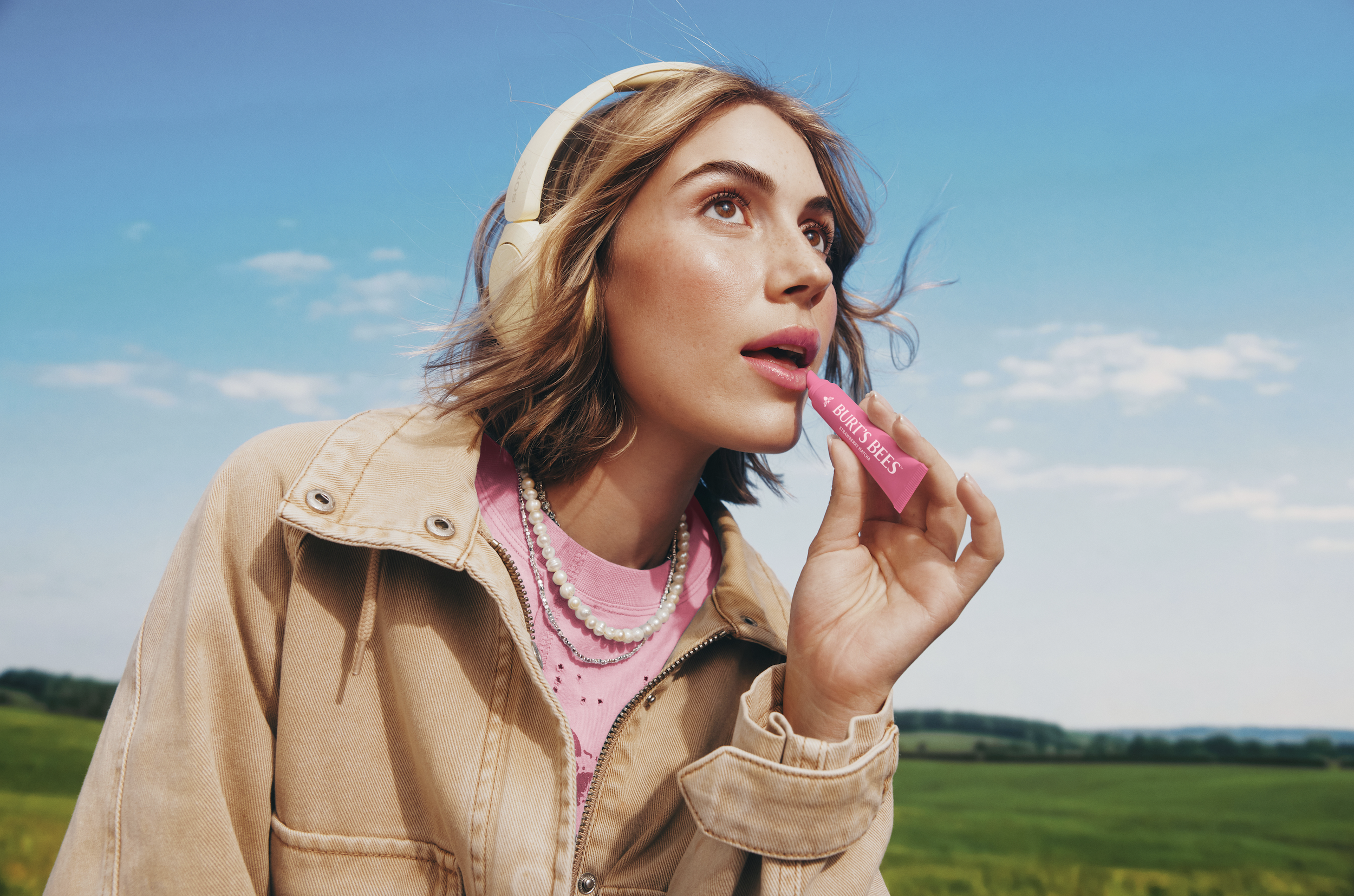 A young woman with brown hair wearing headphones, a beige jacket, a pink shirt, and pearl necklaces, standing outdoors against a blue sky with a few clouds and green fields in the background, applying Burt's Bees lip balm.