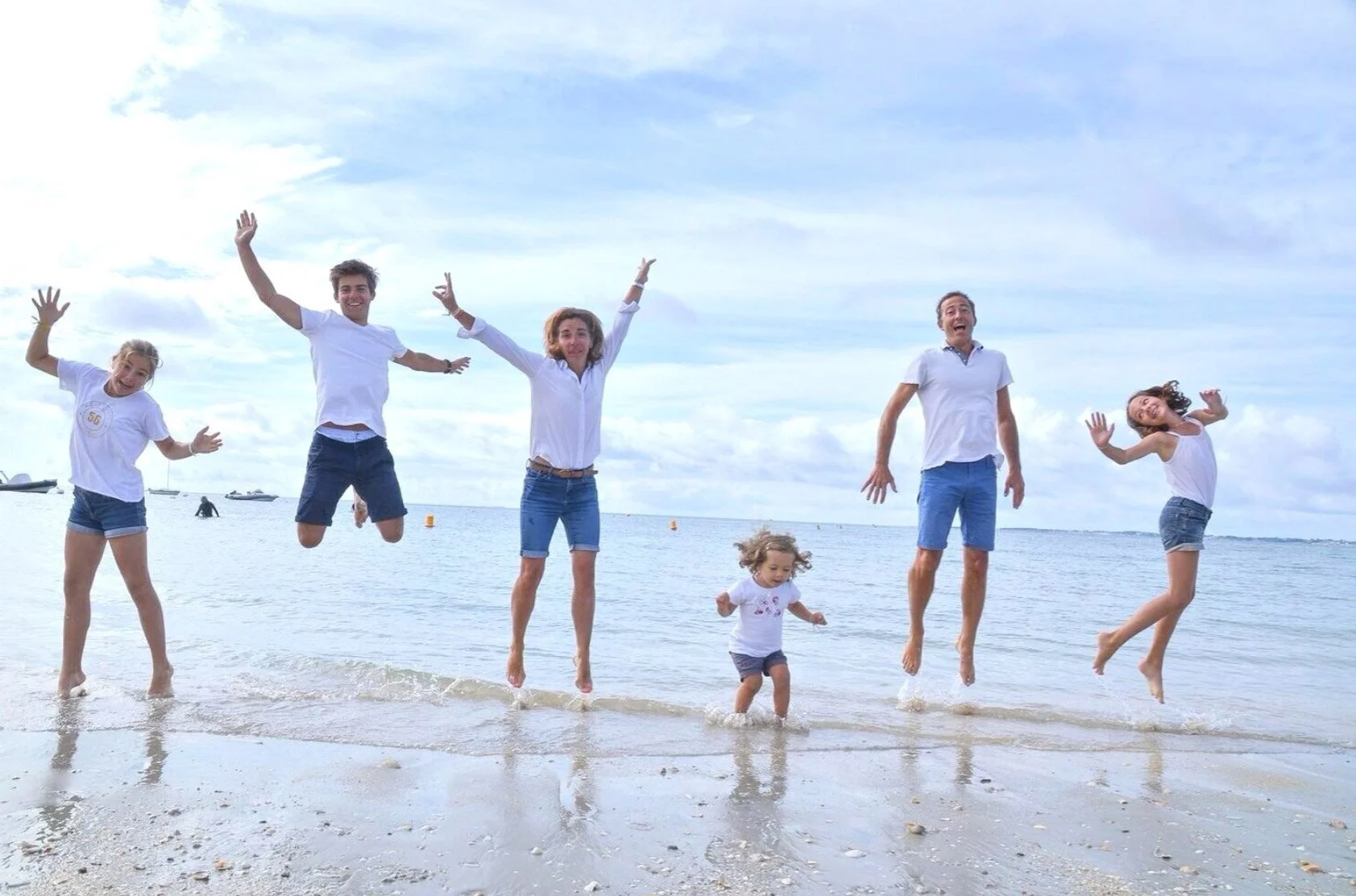 Une famille de six personnes, dont deux enfants, saute en l'air sur la plage au bord de la mer par une journée ensoleillée, souriant et profitant du moment de détente.