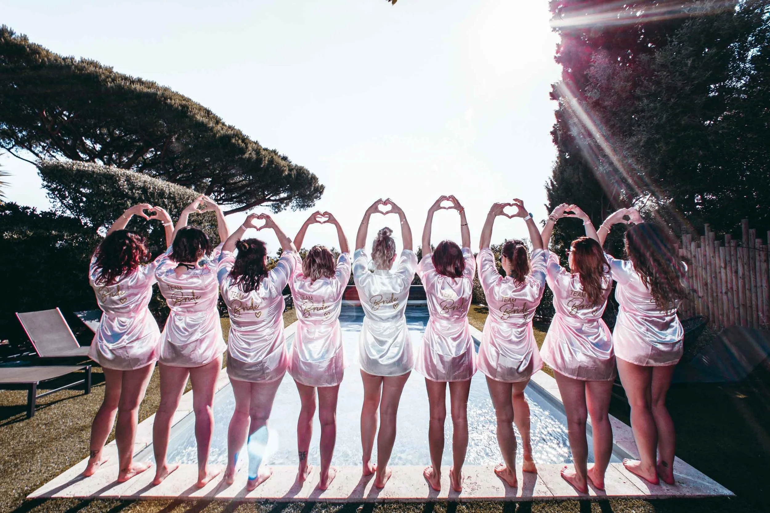 Friends in Team Bride robes making heart shapes with their hands by the pool of a private villa during a bachelorette party in Cavalaire-sur-Mer