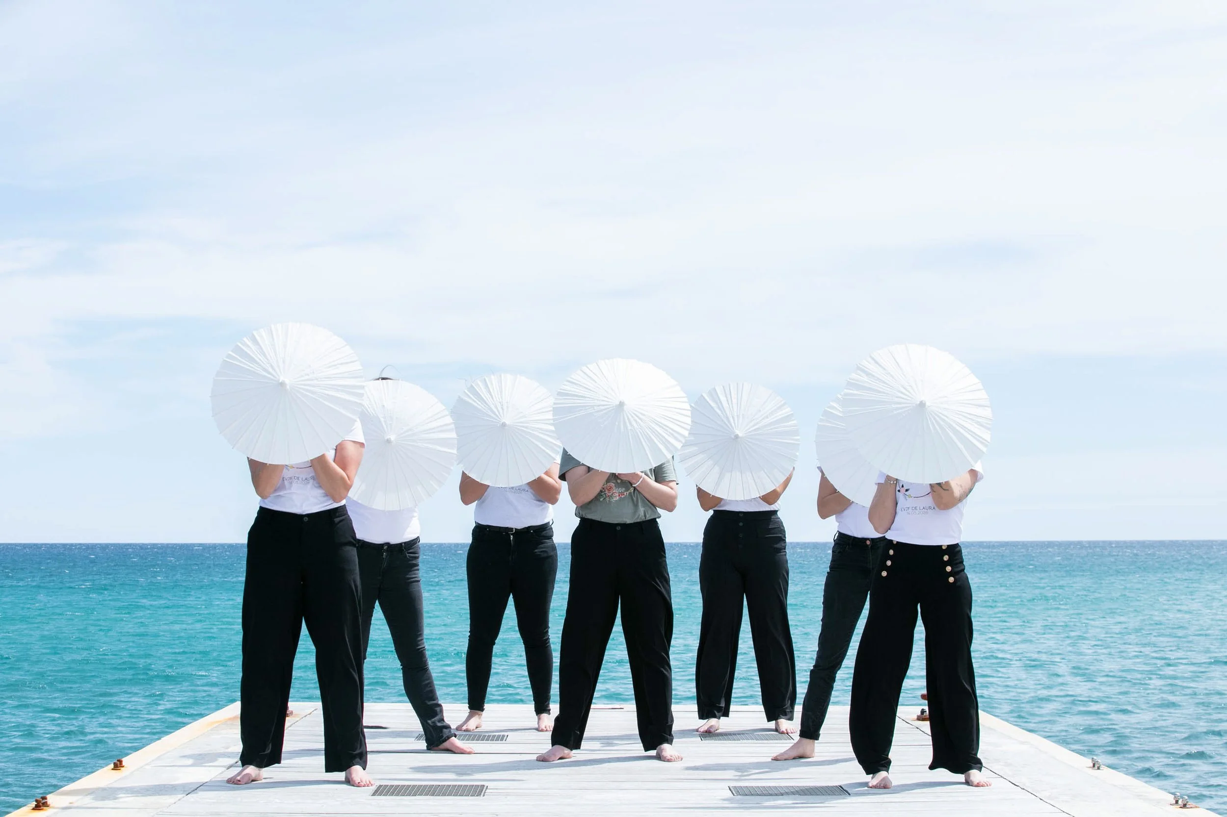 Group of women from behind on a jetty holding white parasols facing the Mediterranean Sea during a hen party photo session on the French Riviera