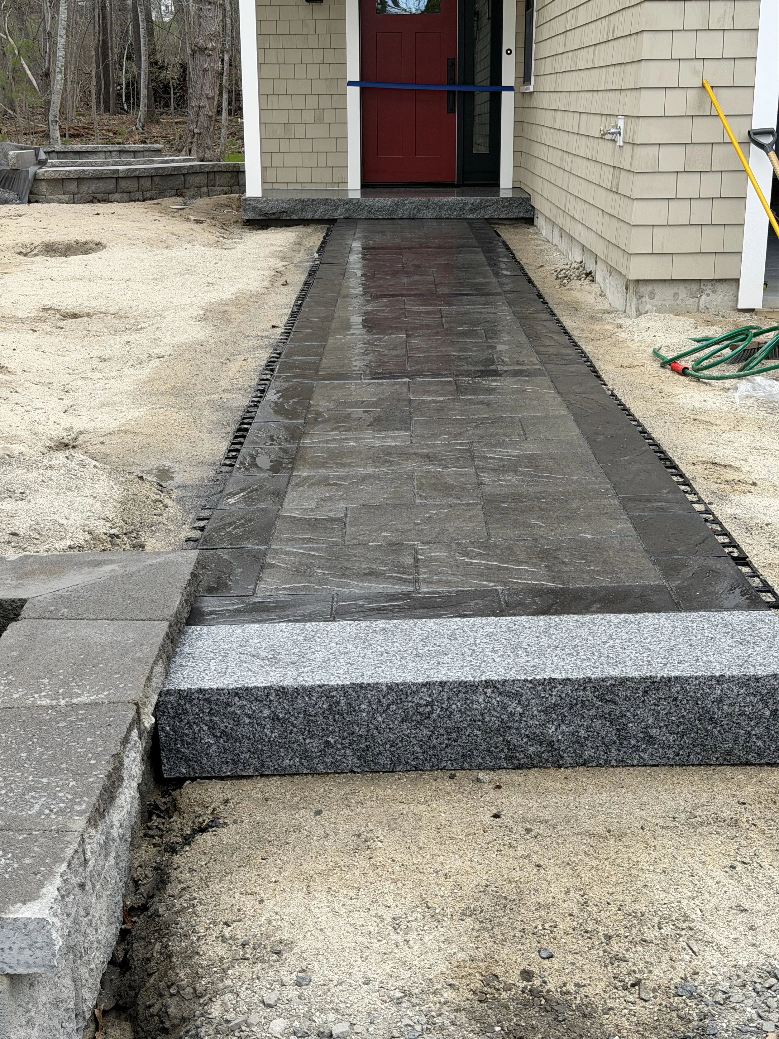 Freshly laid stone pathway leading up to the front door of a house. The path is bordered by a black drainage grate and surrounded by dirt and gravel. A red front door is visible in the background, with a blue tape across the doorframe.