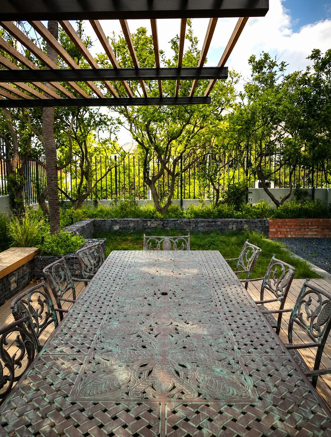 Outdoor patio with a large metal table and eight chairs, surrounded by trees and greenery, with a wooden pergola overhead.