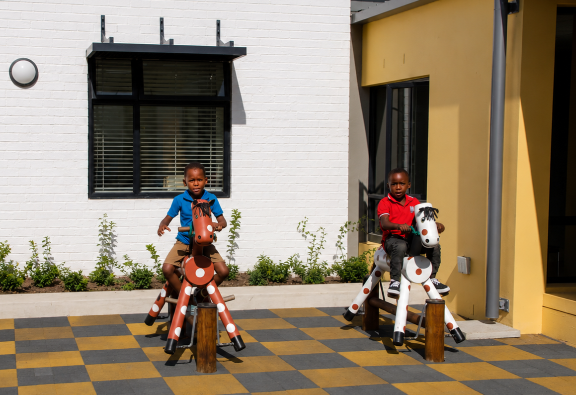 Two children riding on animal-shaped playground toys and a third child walking nearby on a checkered yellow and gray surface in a sunny outdoor area with modern buildings in the background.