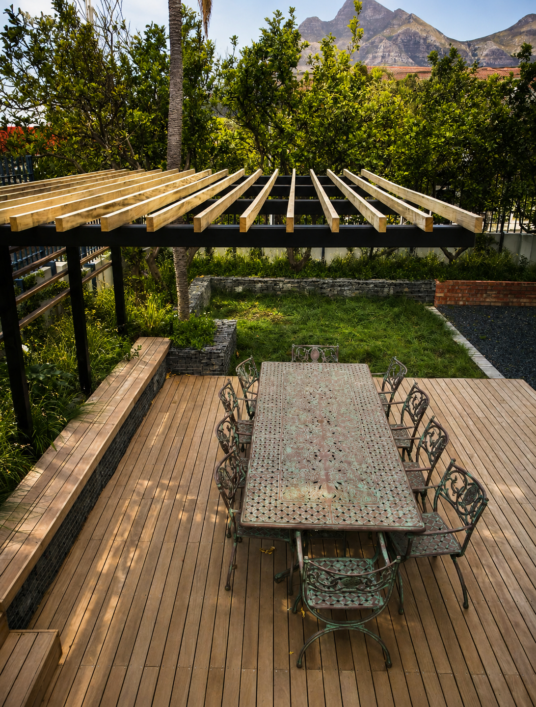 Outdoor dining area with a metal table and chairs on a wooden deck, adjacent to a grassy backyard with trees, a brick wall, and a mountain view in the background.