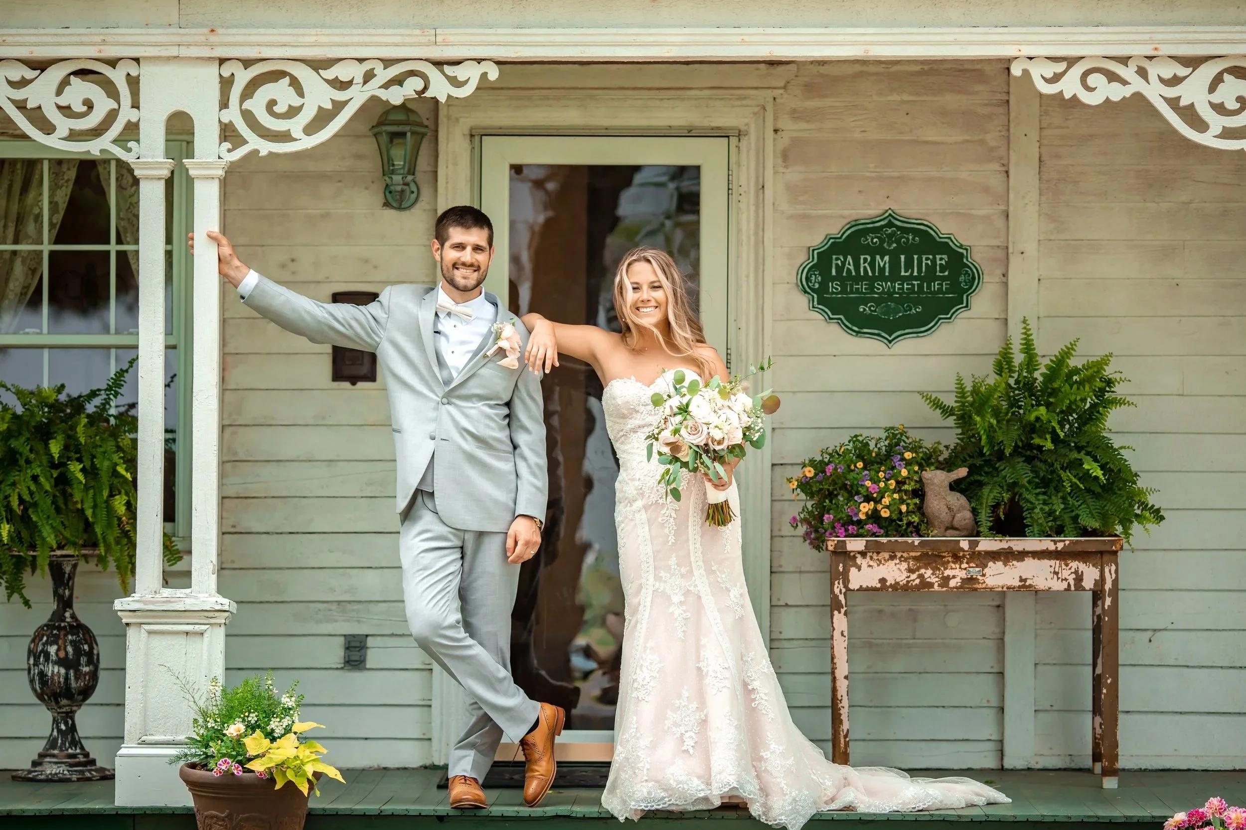 A smiling bride in a lace wedding gown holding a bouquet of flowers, standing next to a groom in a light gray suit with a white bow tie, outside a rustic wedding venue in Illinois with decorative plants and a sign that reads 'Farm Life is the Sweet L