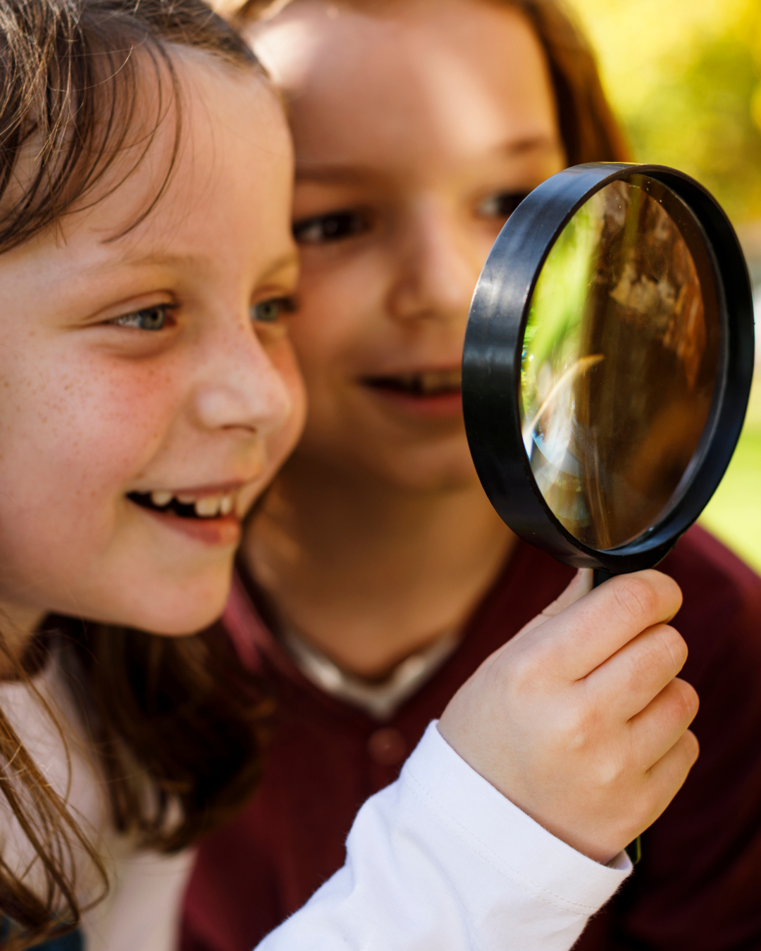 Two children looking through a magnifying glass outdoors, observing something closely with interest and curiosity.