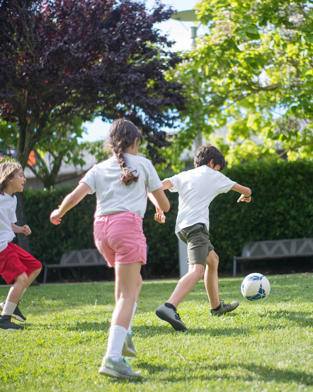 Children playing soccer outdoors on a sunny day with trees and benches in the background.