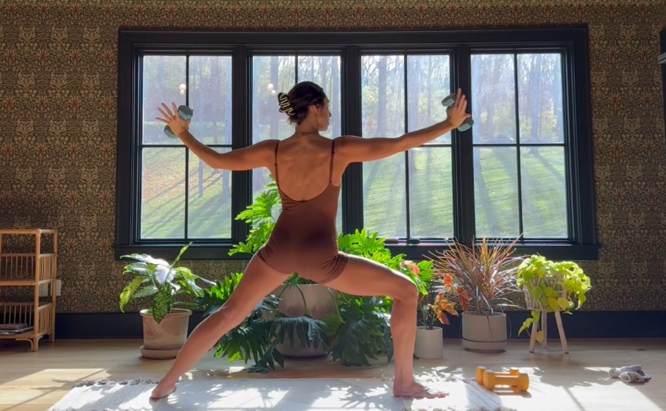 A person standing on a yoga mat in front of large windows with a view of a green outdoor landscape, dressed in workout attire.