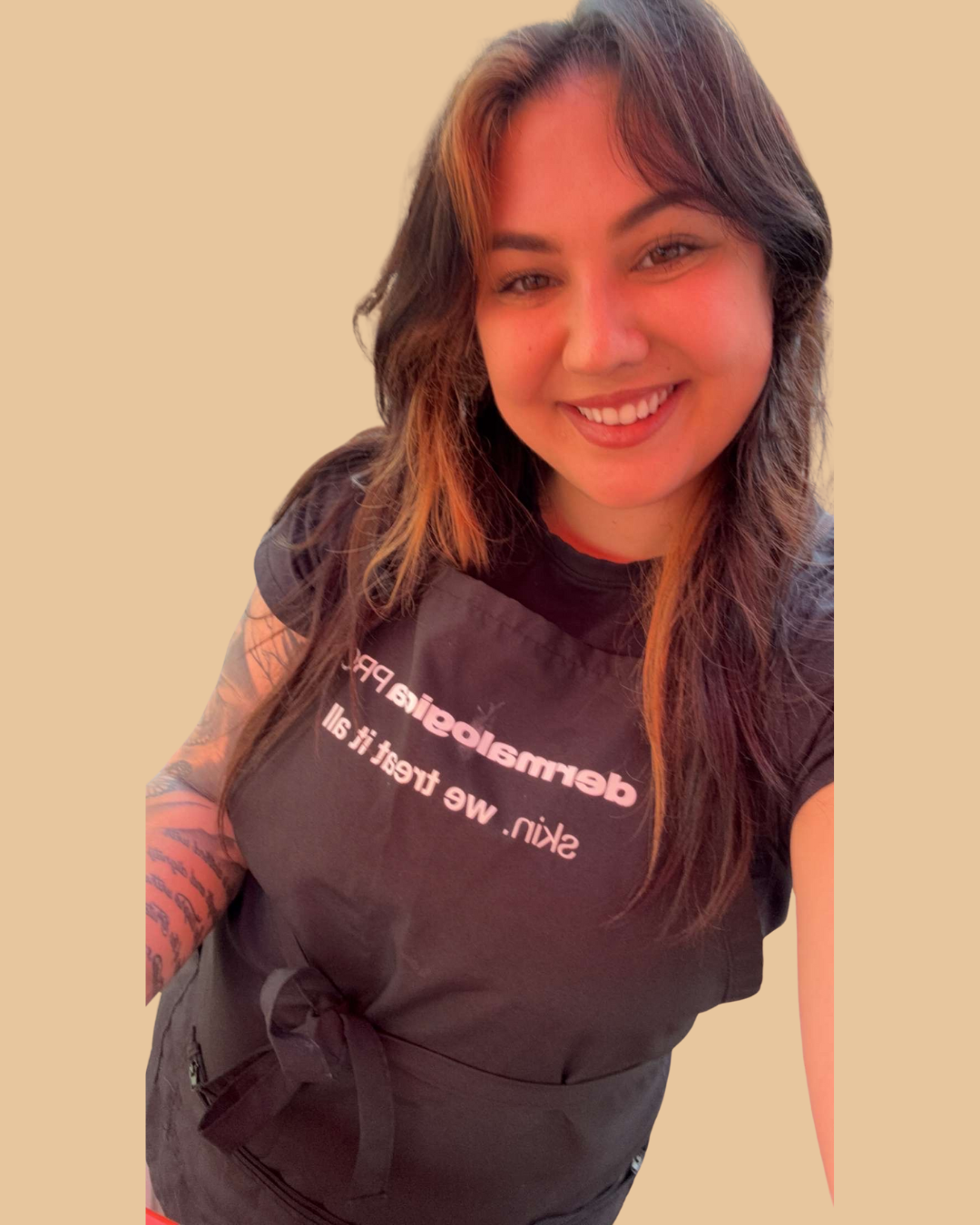 A woman with long brown hair smiling, wearing a black apron with the words 'demmelogistics PR' and 'skl. we treat it all' printed on it, against a plain beige background.
