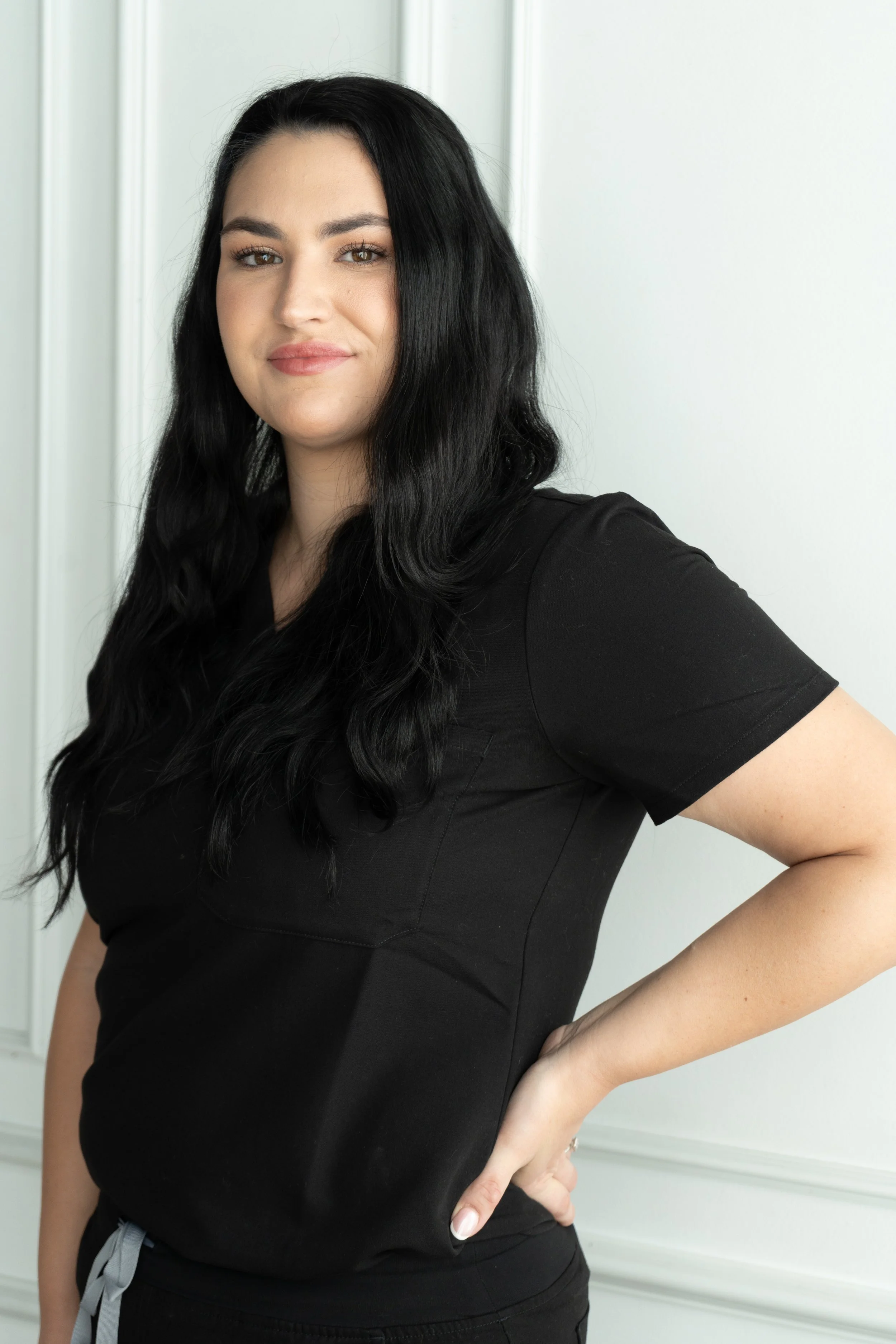 A woman with dark hair, light skin, and makeup, smiling at the camera, wearing a white top and a dark blazer, sitting against a beige background.