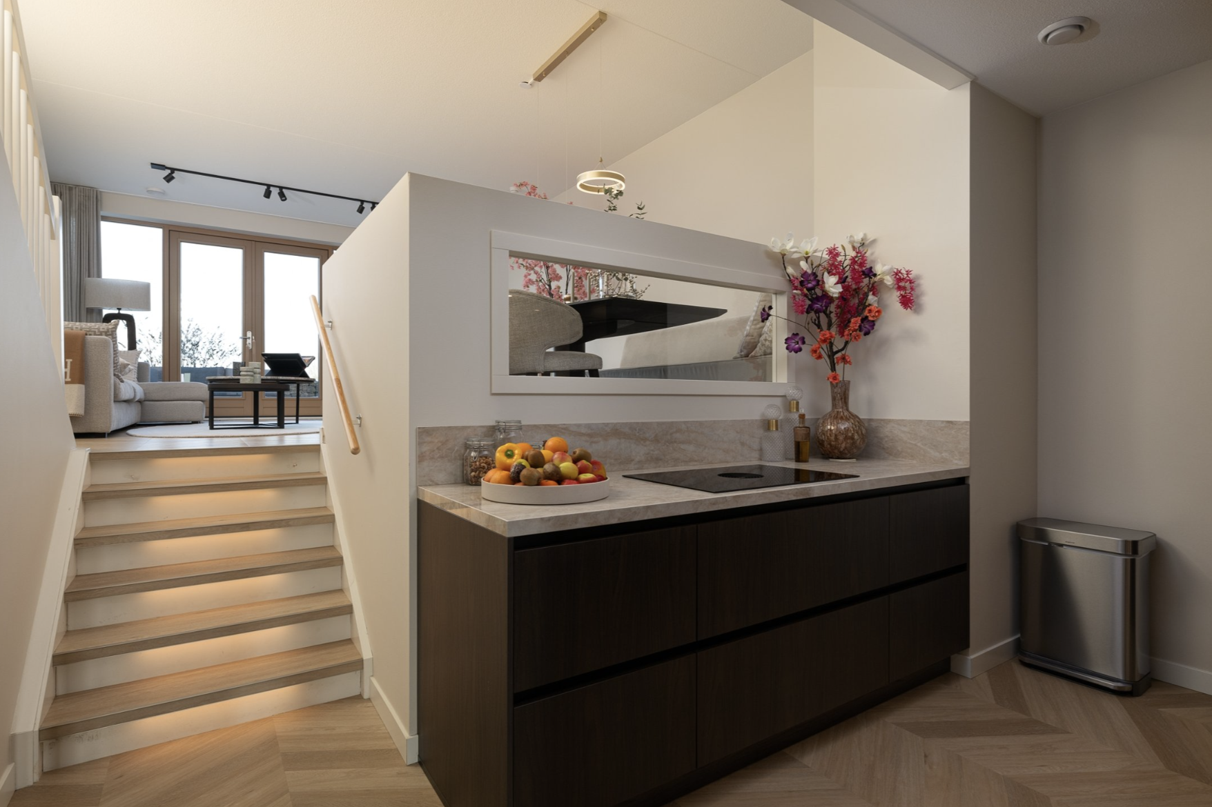 Zwart keukenblok met fruit en bloemen, trap naar woonkamer met grote ramen en zithoek.