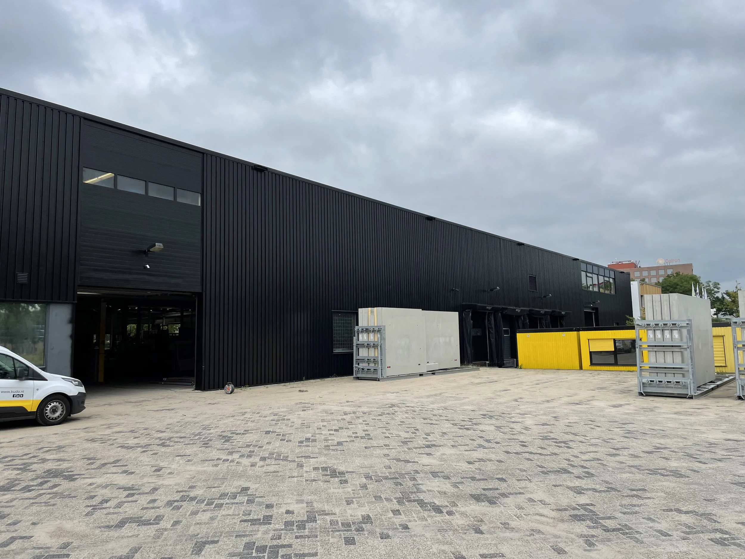 Nieuw, modern zwart industrieel gebouw onder een bewolkte hemel, met enkele gele en grijze constructies en een auto op een bestrate parkeerplaats.