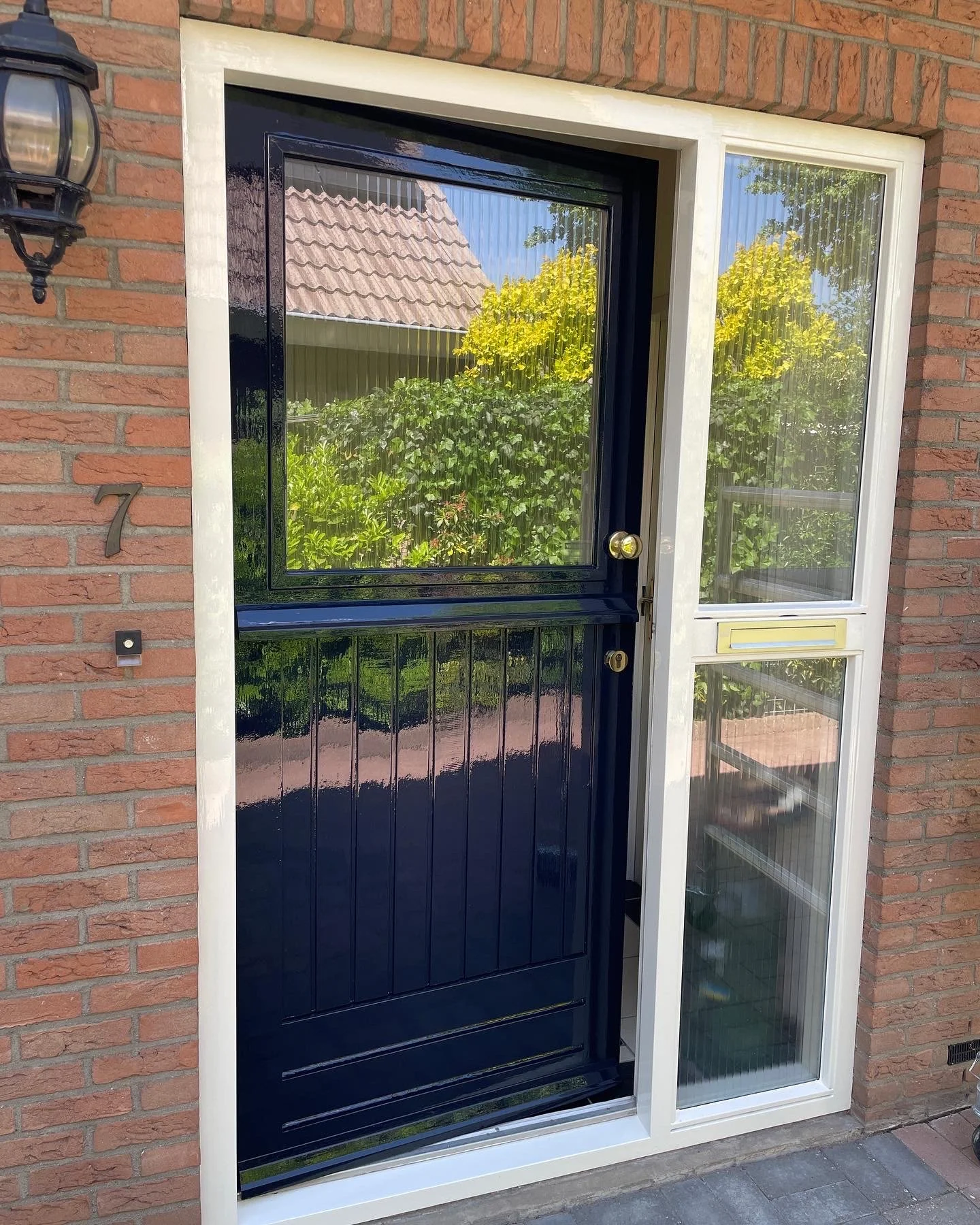Gezicht op een voortuin met een zwarte deuropening en een bijpassende deur, geflankeerd door een wit frame en een raam met pigmentglas, met een groene tuin en een huis met een tegeldak zichtbaar door het raam
