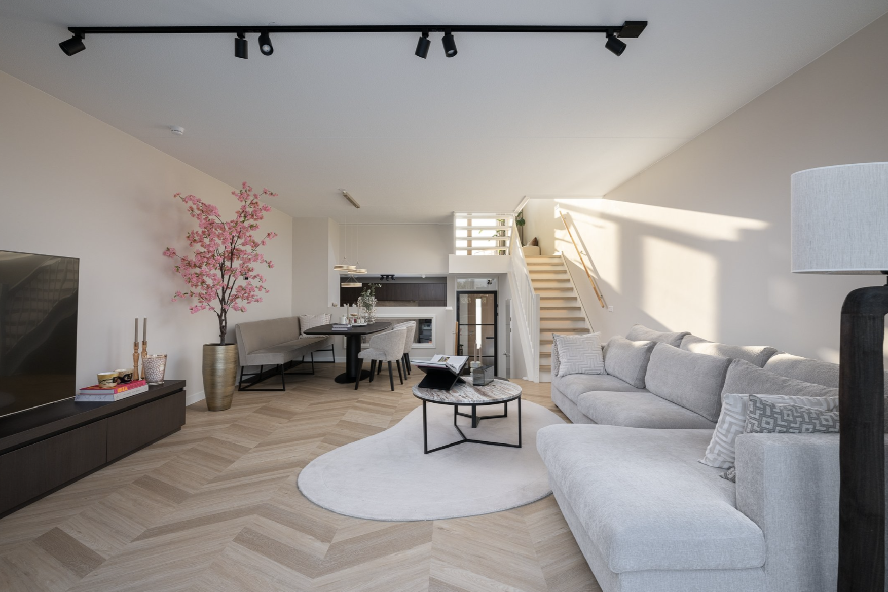 Licht woonkamer met grote lichte bank, bloemen op tafel, televisie, en eettafel met stoelen