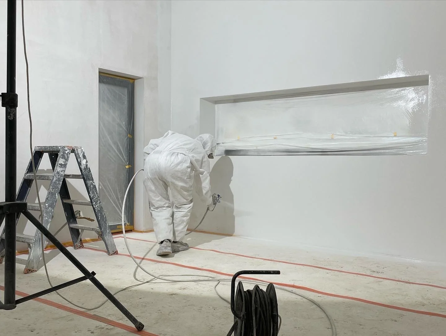 Een persoon in all white beschermkleding spuit verf op een witte muur in een kamer. Er is een ladder en een elektrische kabel op de vloer en een raam dat afgeplakt is.