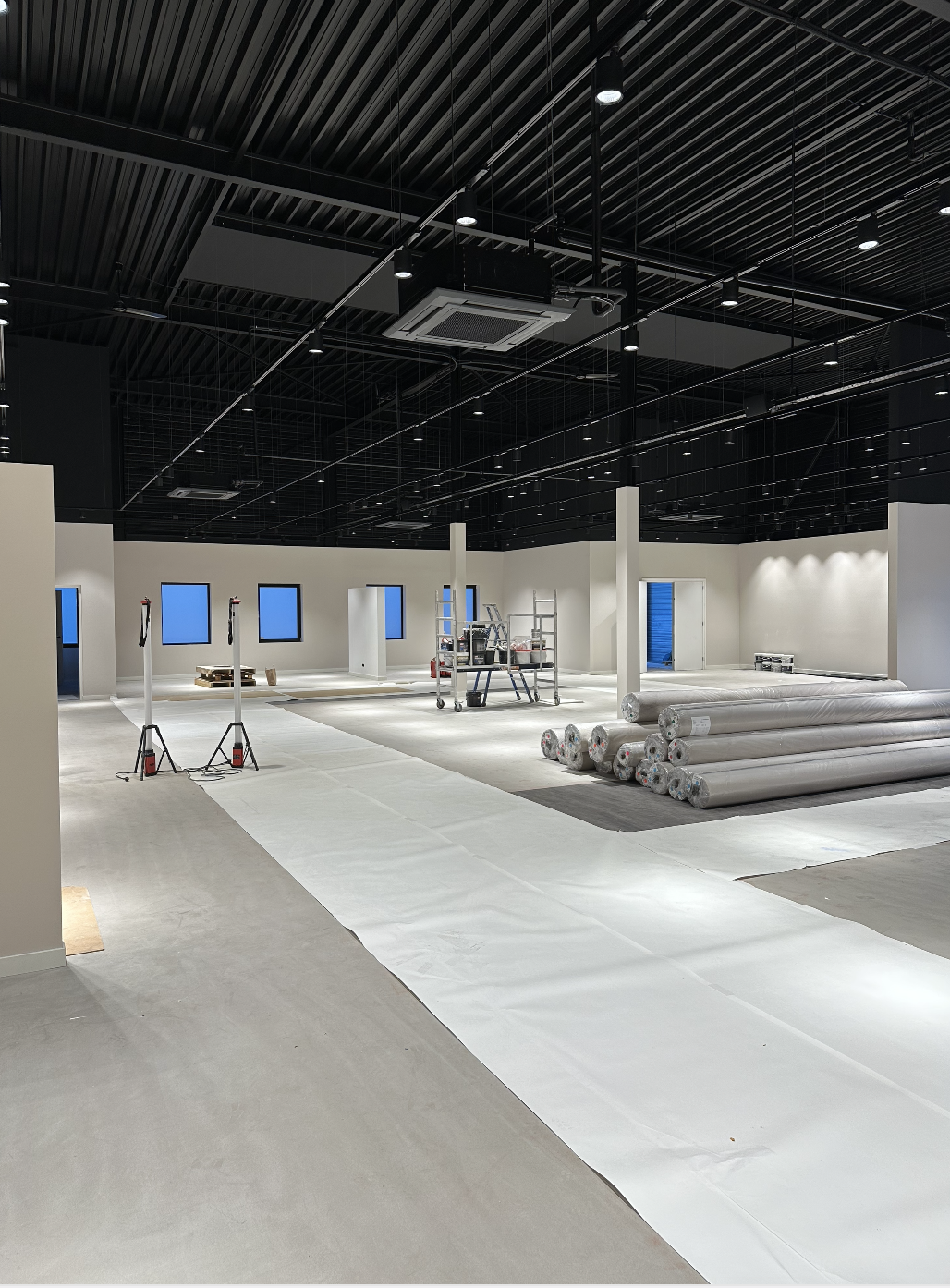 Interieur van een winkel of kantoorruimte onder constructie, met vloerbedekking, plafondlichten, en bouwmaterialen zoals rollen tapijt en gereedschap.