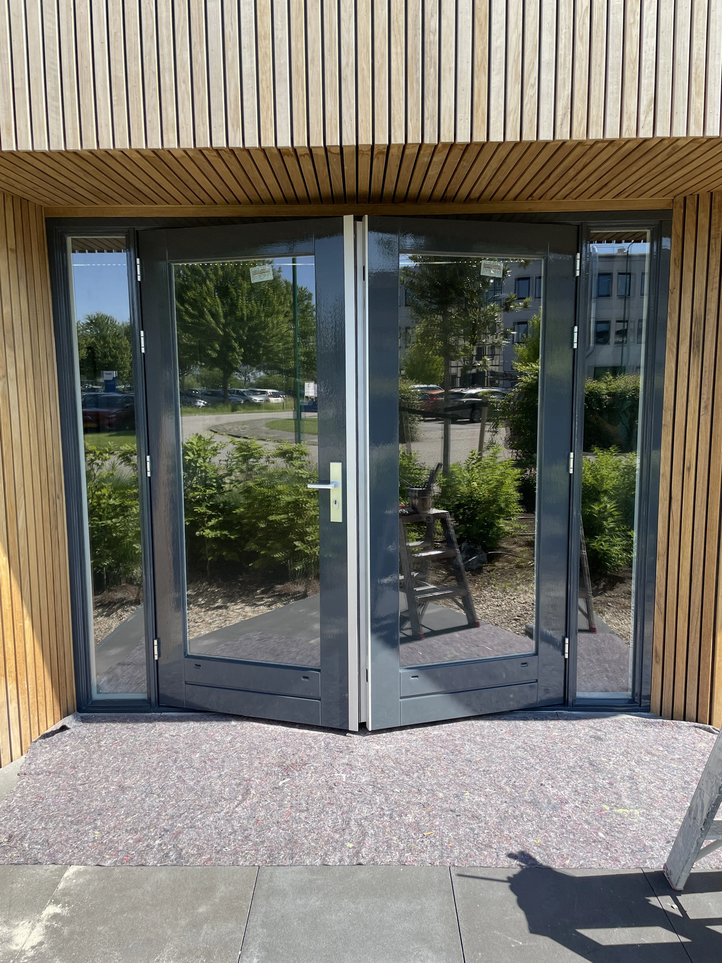 Modern glazen deuropening met houten gevelbekleding en een mattenmat voor de ingang, met een tuin en een parkeerplaats op de achtergrond.