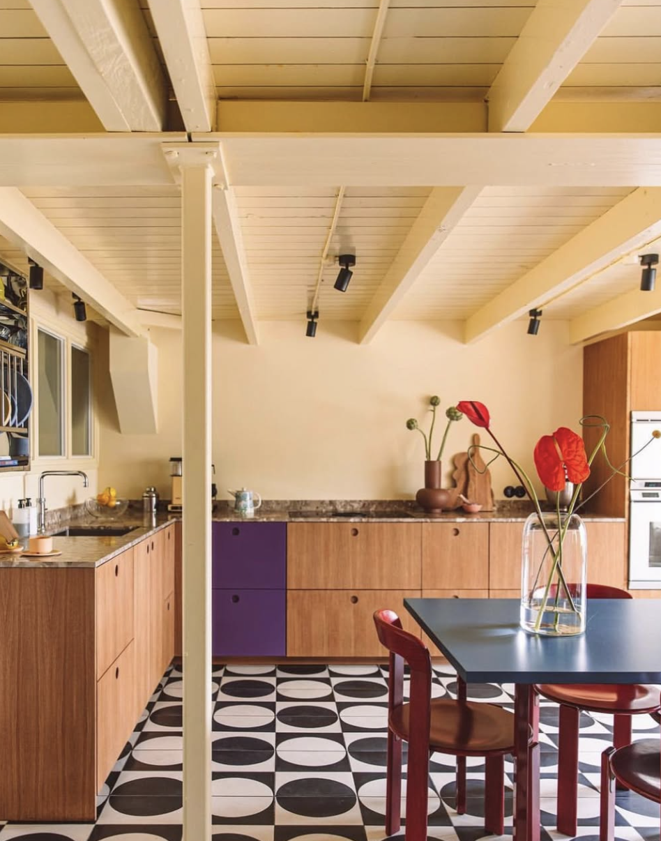 Keuken met houten kasten, geel geschilderde wanden, plafond met houten balken en zwarte spotlights.