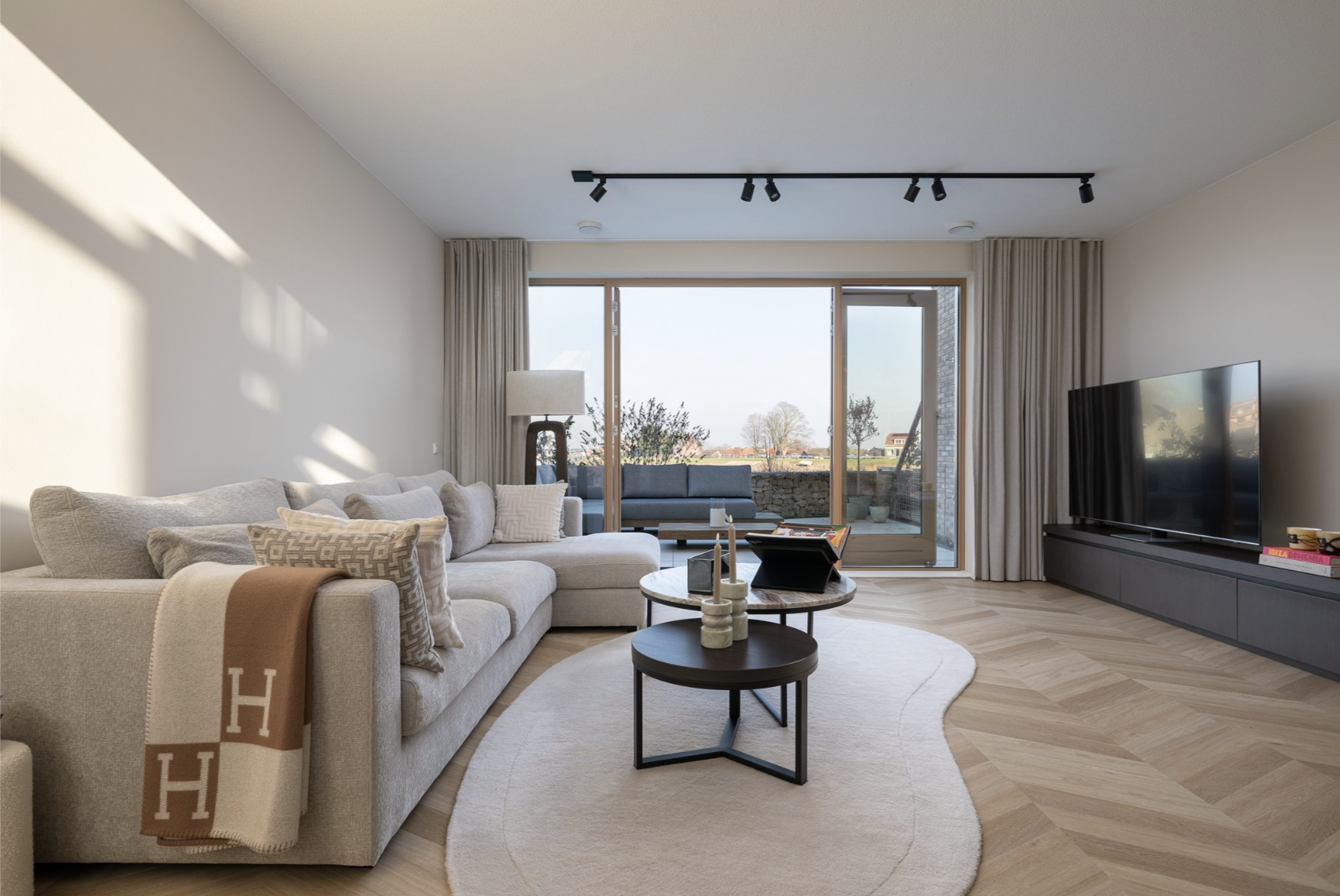 Modern woonkamer met grote bank, salontafels, tv, en uitzicht op tuin en landschap via grote glazen deuren.