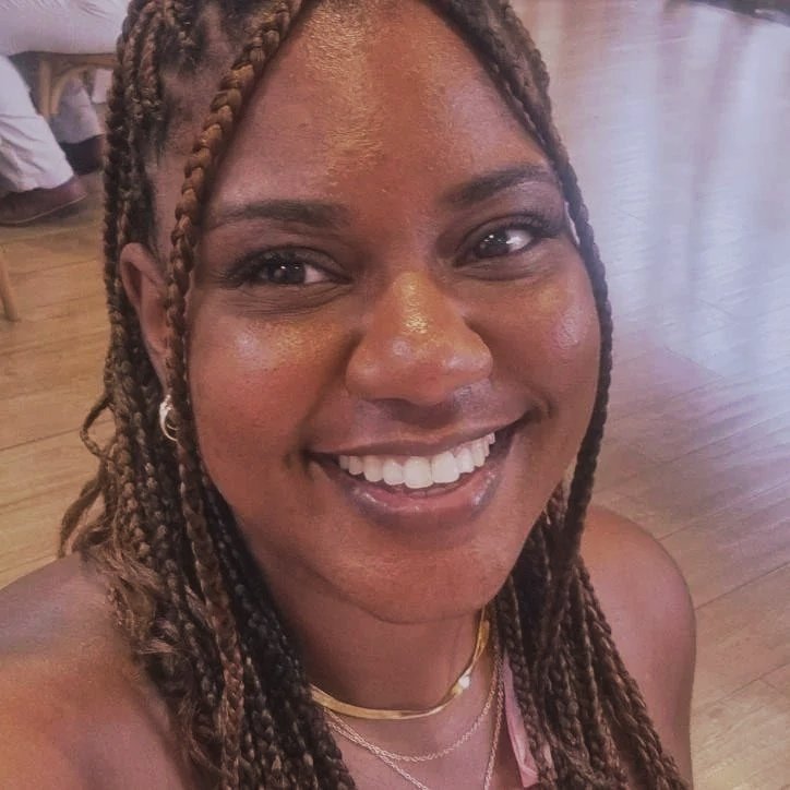 Close-up of a smiling woman with braided hair in a wooden-floored room.