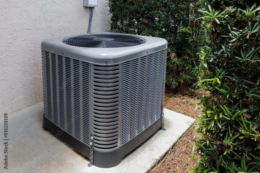 Outdoor air conditioning unit on concrete pad next to house and green bushes.