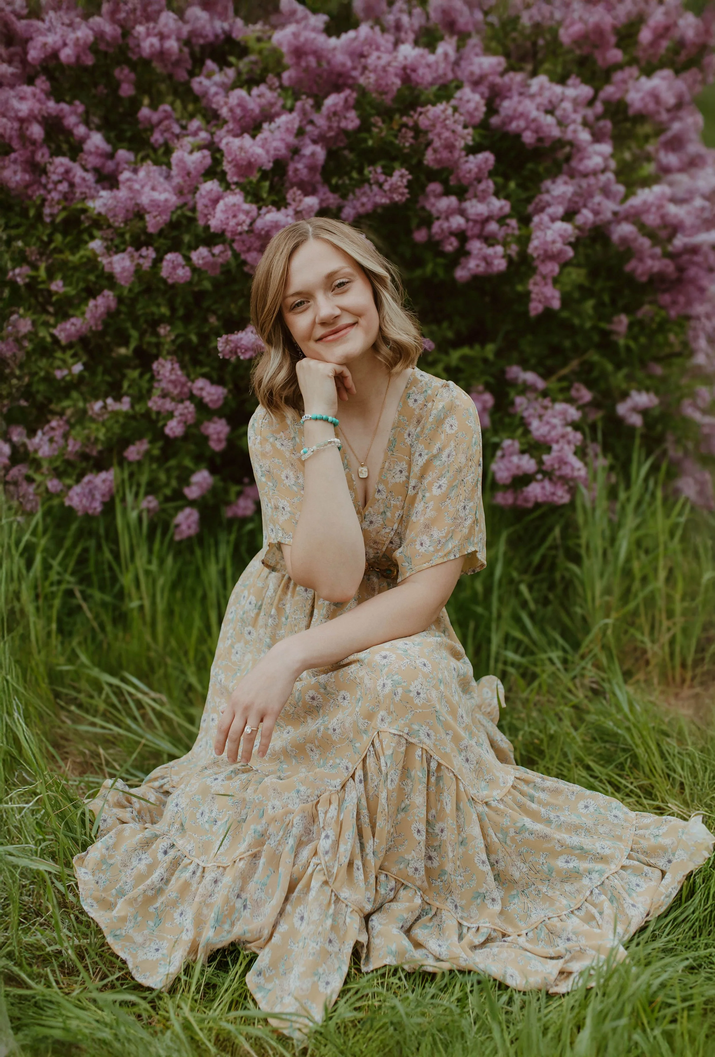 A young woman sitting on the grass in front of a flowering bush with pink blooms, wearing a flowy floral dress and jewelry, smiling gently.