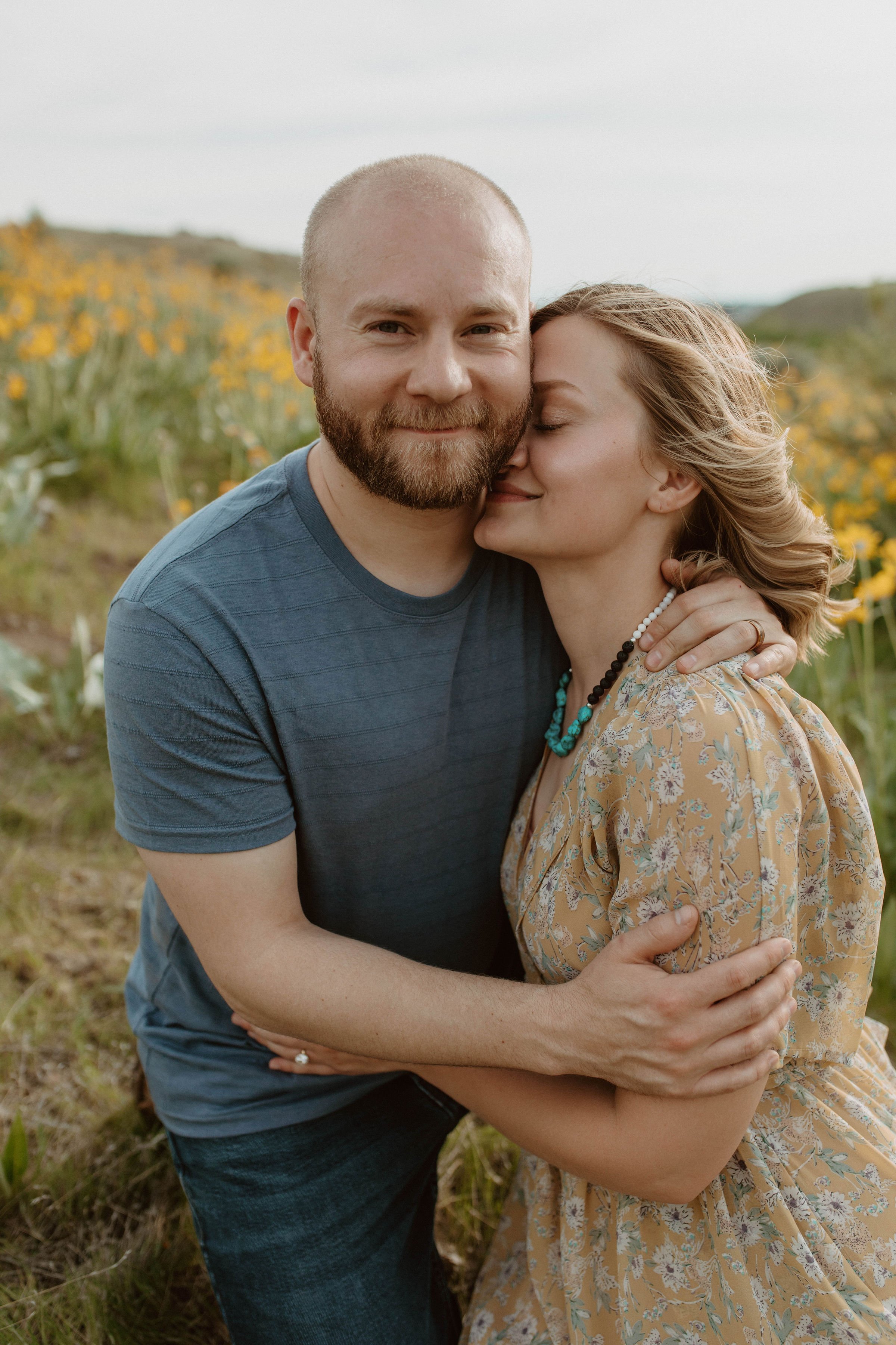 A couple embracing outdoors in a field of yellow flowers, smiling gently.