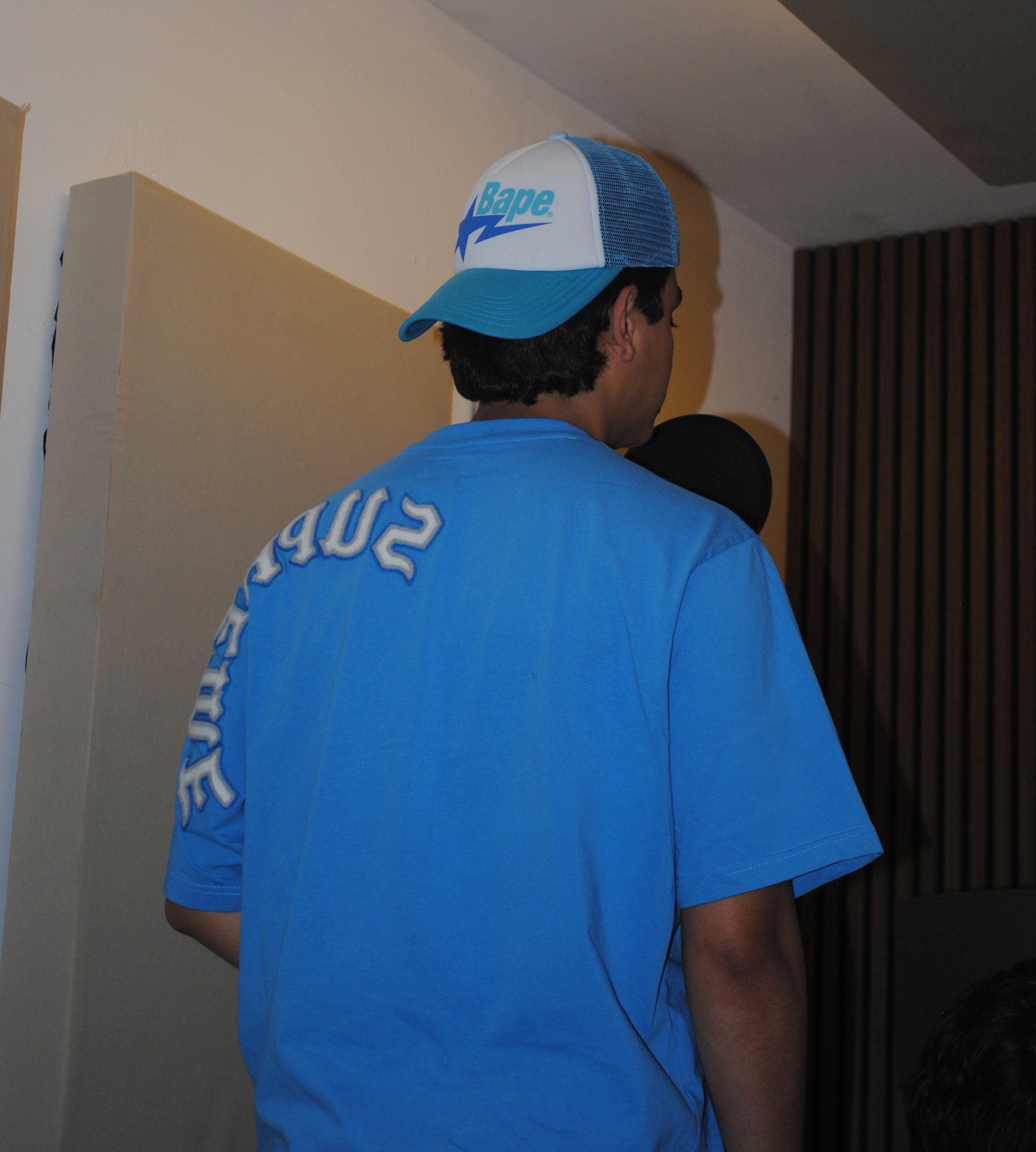 Hombre de espaldas con camiseta azul clara y gorra blanca y azul con la marca Bape, en una habitación con paredes blancas y una pared de madera oscura
