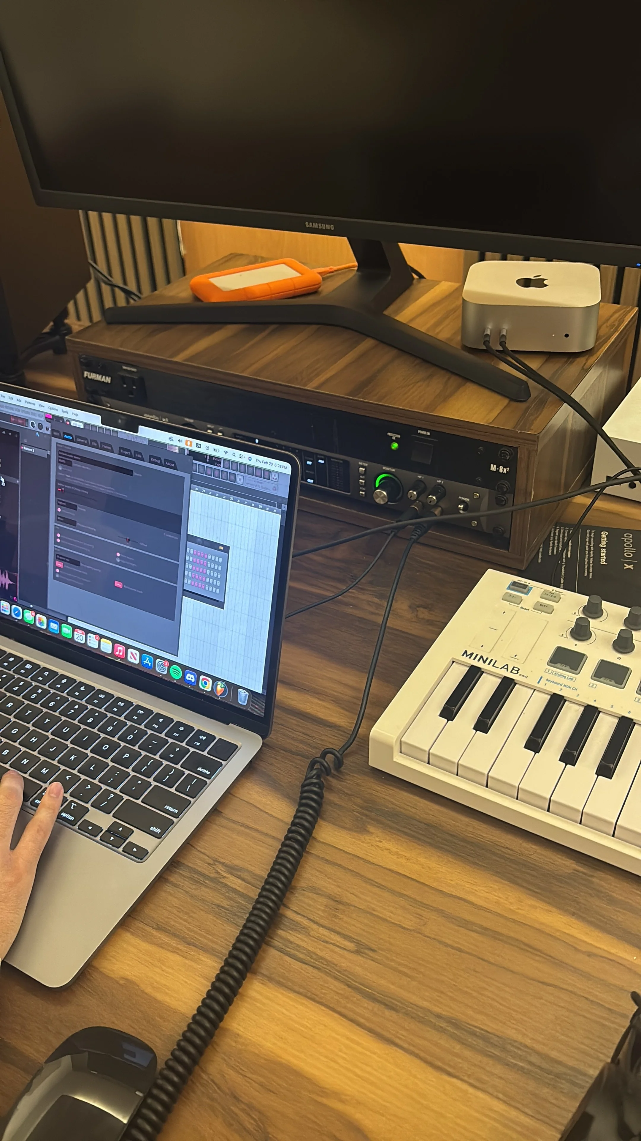 Estación de trabajo de producción musical con una computadora portátil, un controlador MIDI, un monitor, una caja de almacenamiento y un disco duro externo en una mesa de madera.