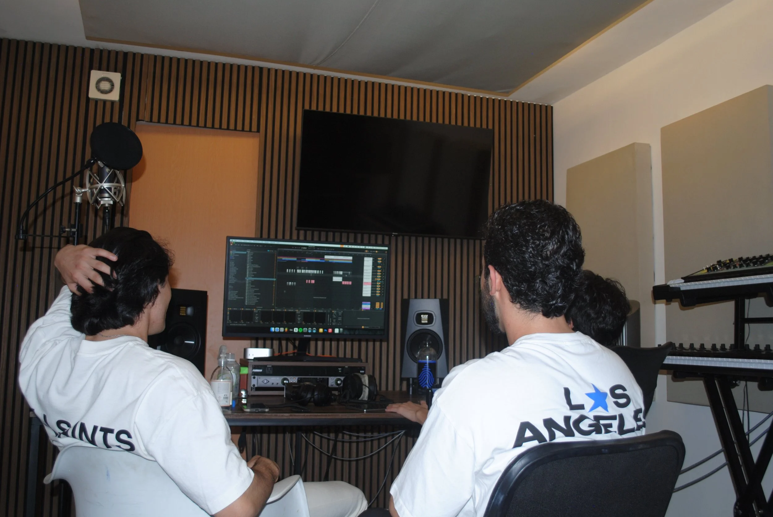 Tres personas en un estudio de grabación, uno trabaja en una computadora con un software de edición de audio, y las otras dos observan. Están usando camisetas blancas con el texto 'LOS ANGELES'.